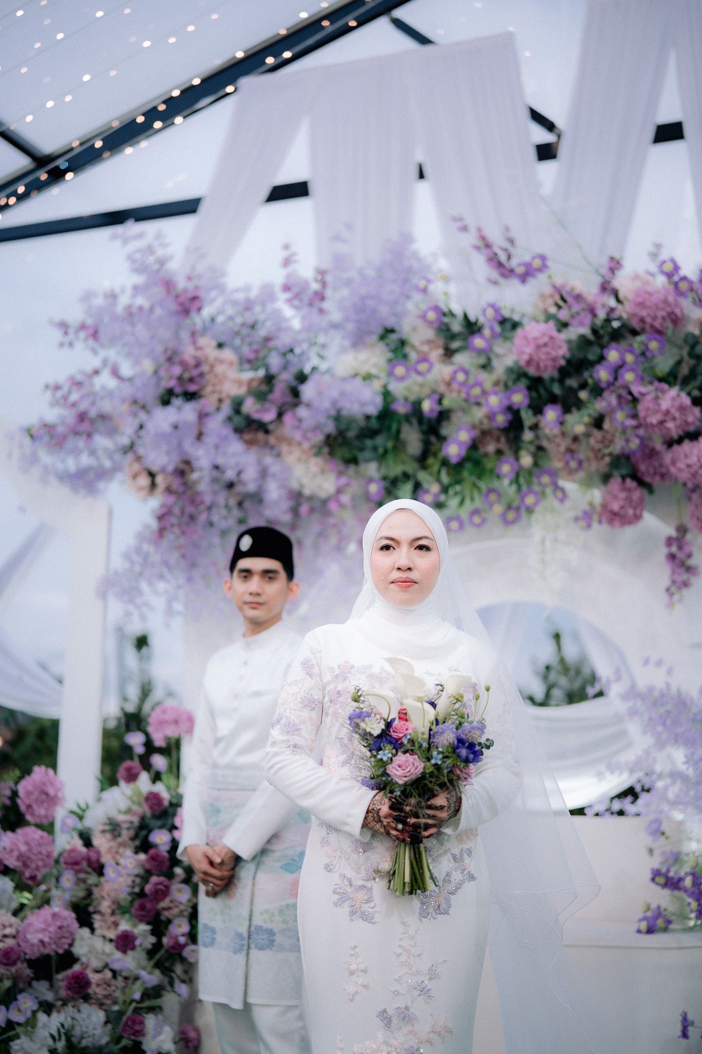 Malay couple in traditional white wedding attire pose among purple floral arch decorations at elegant ceremony.