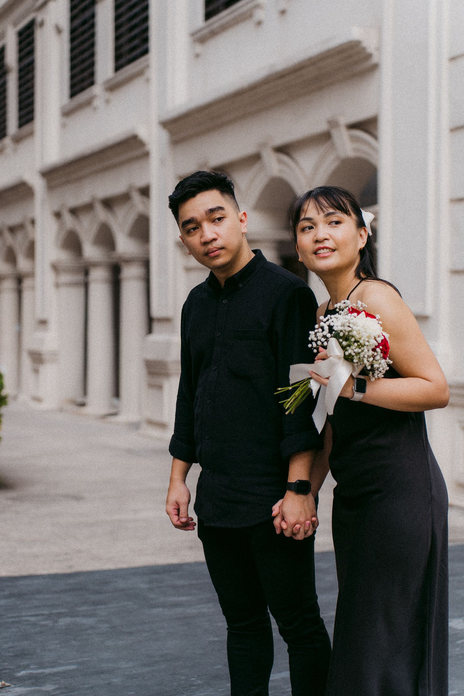 A couple in black formal wear posing together against a white colonial-style building backdrop.
