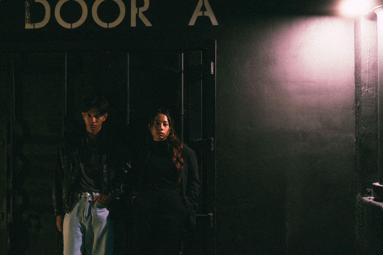 Dark moody shot of figures standing by a door marked 'DOOR A' under dim lighting.