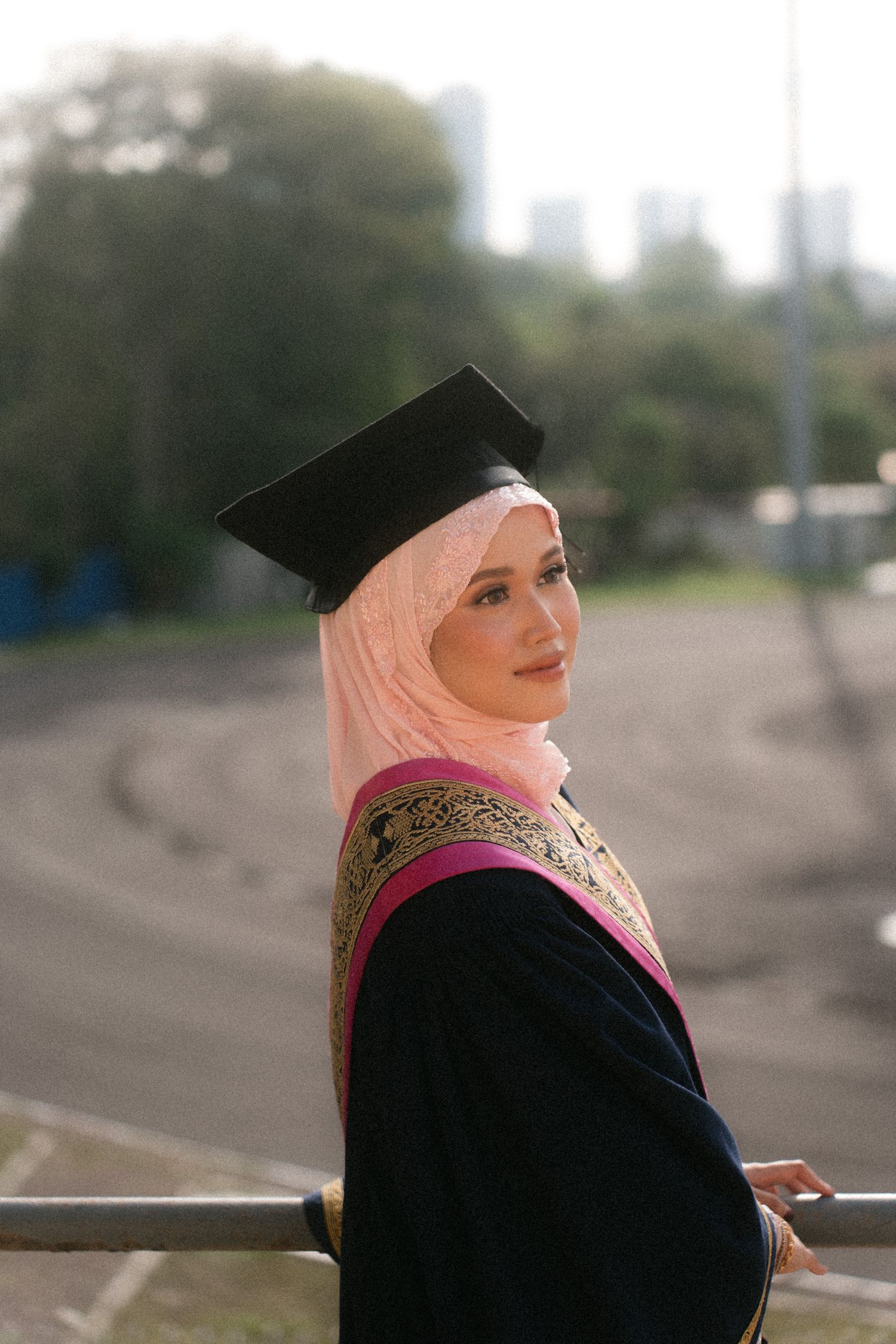 A person wearing a pink hijab and black clothing looks to the side while seated outdoors.