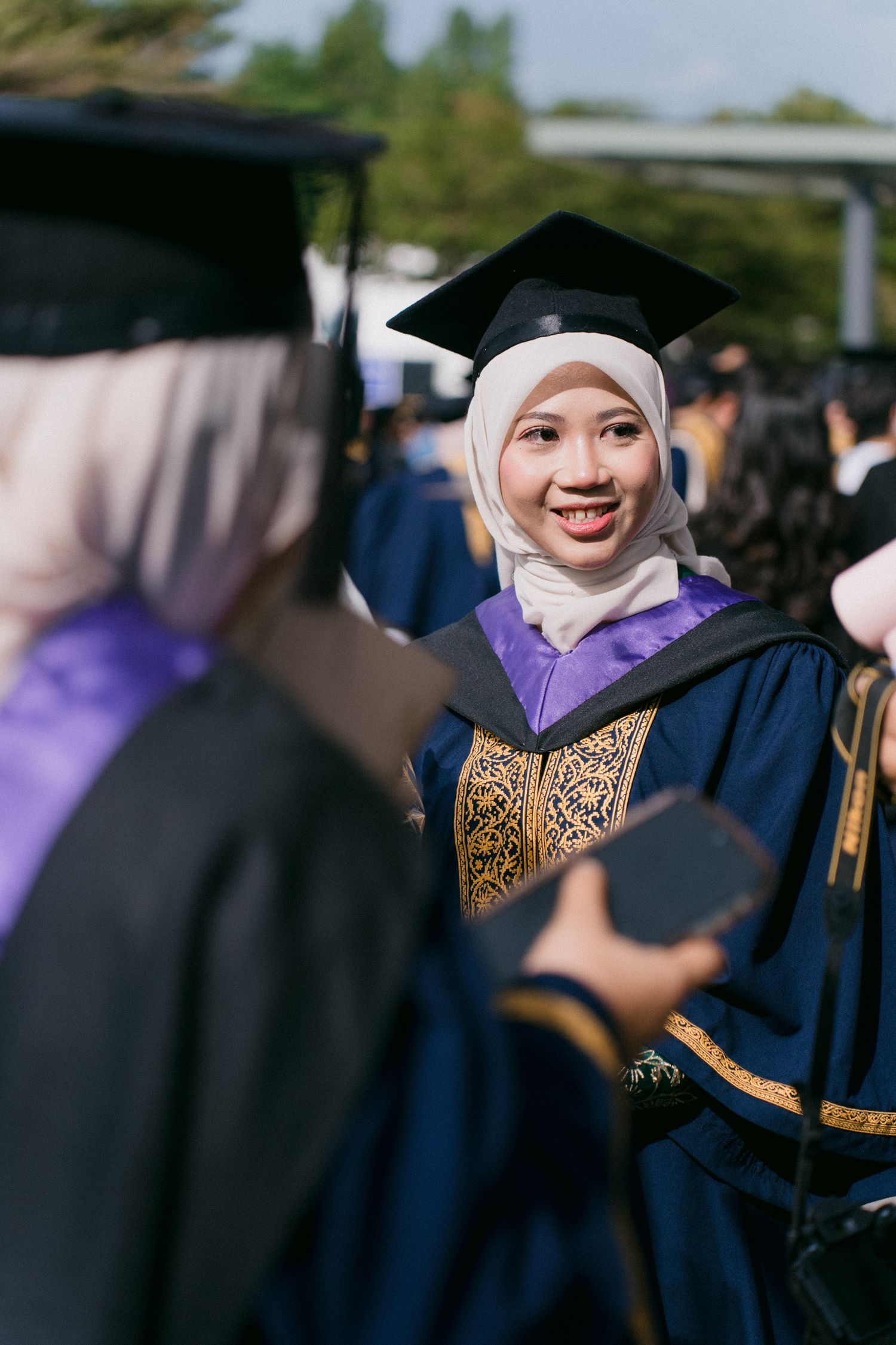 Graduates in dark blue academic regalia with purple accents celebrate during a university commencement ceremony.