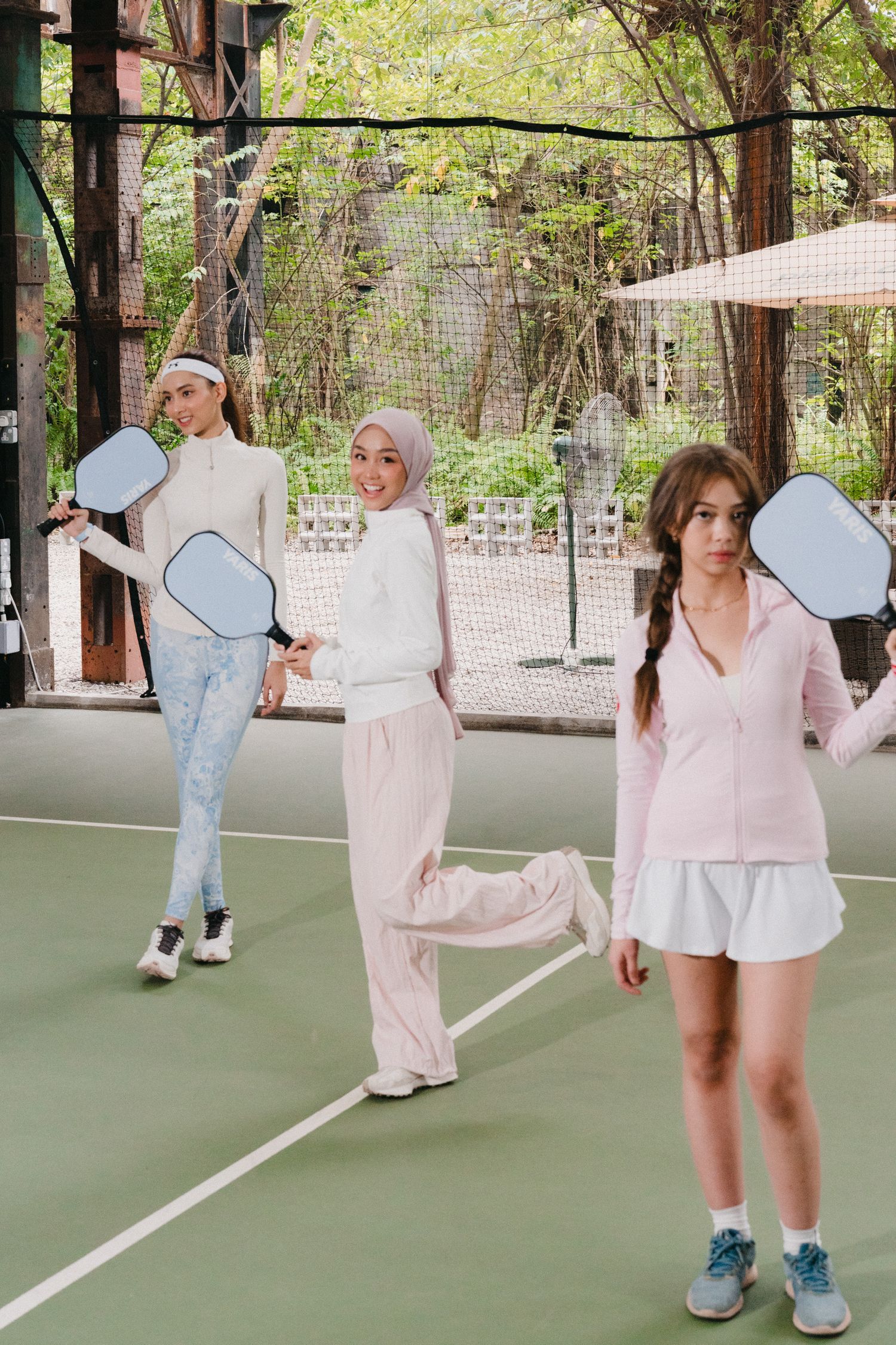 Two people in casual athleisure outfits play tennis on an outdoor court surrounded by trees.