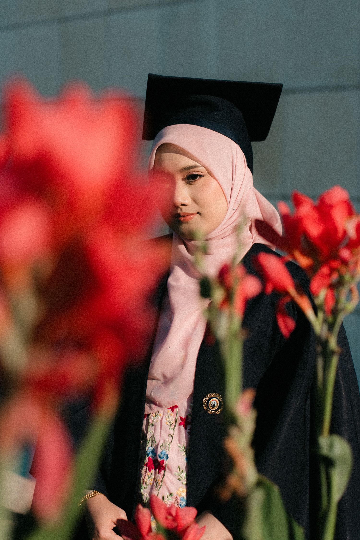 Graduate in pink hijab and floral dress poses with red flowers in a graduation photoshoot.