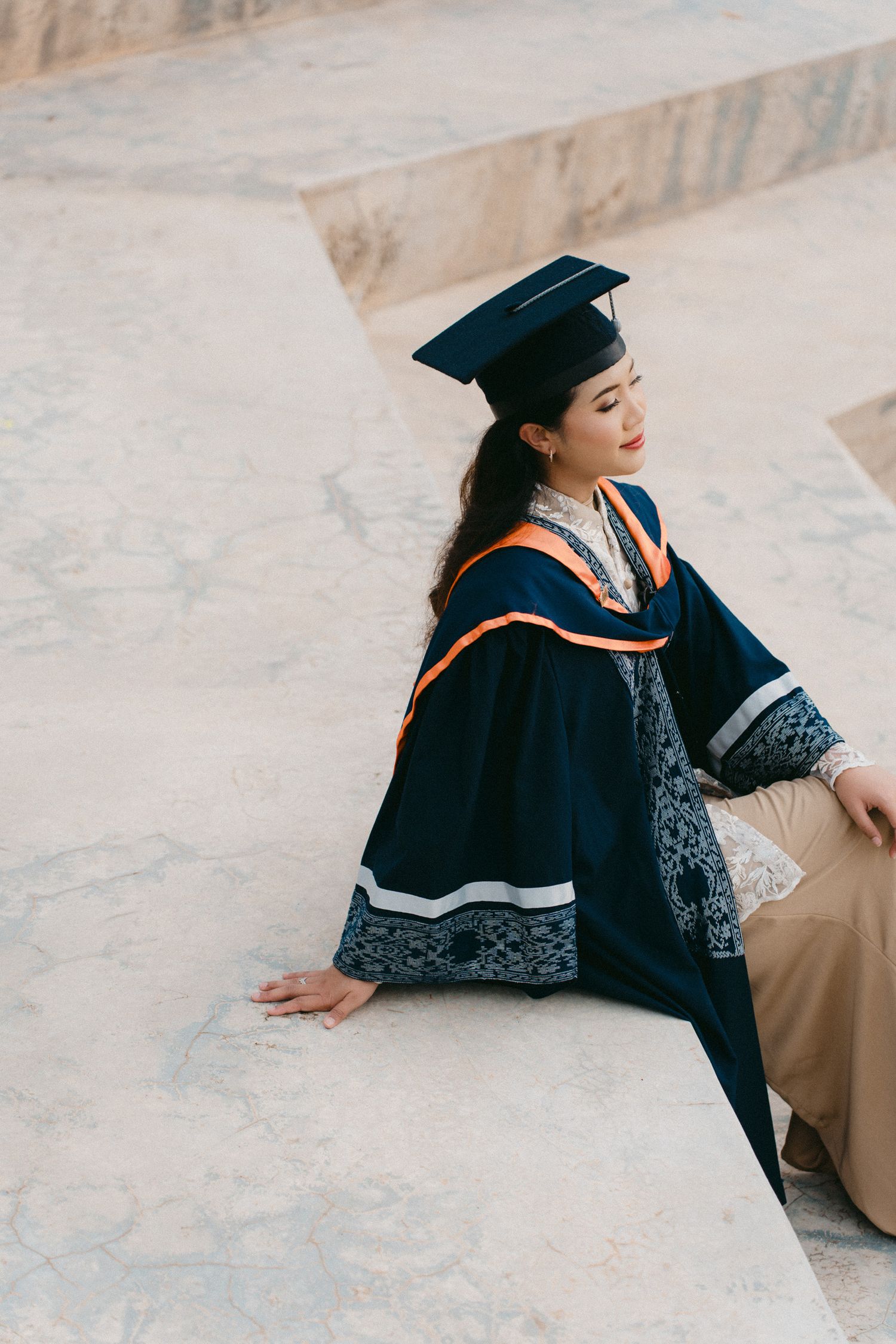 Graduate in black cap and gown sits casually on stone steps during graduation photoshoot.