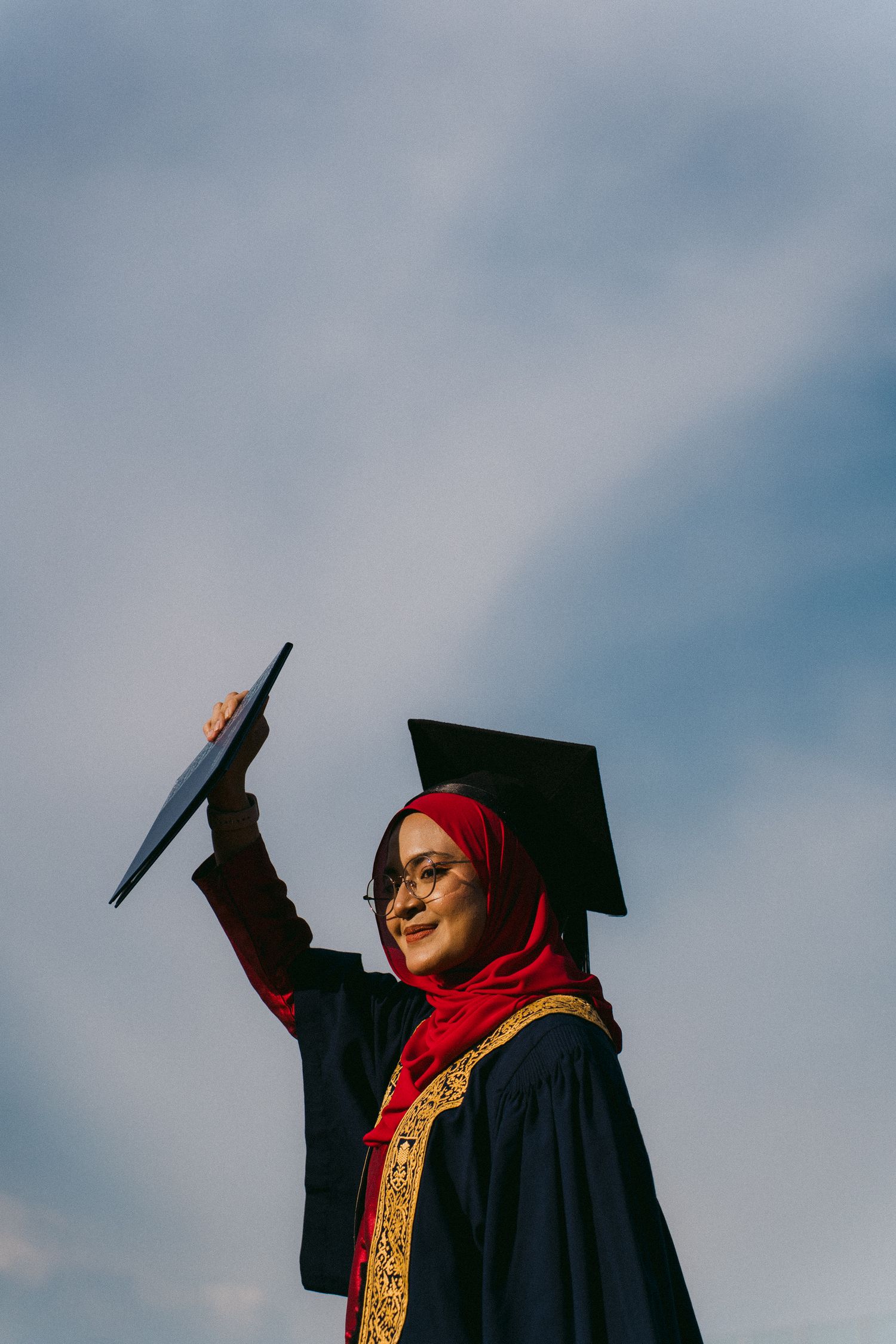 Graduate in traditional academic dress raises cap triumphantly against blue sky.
