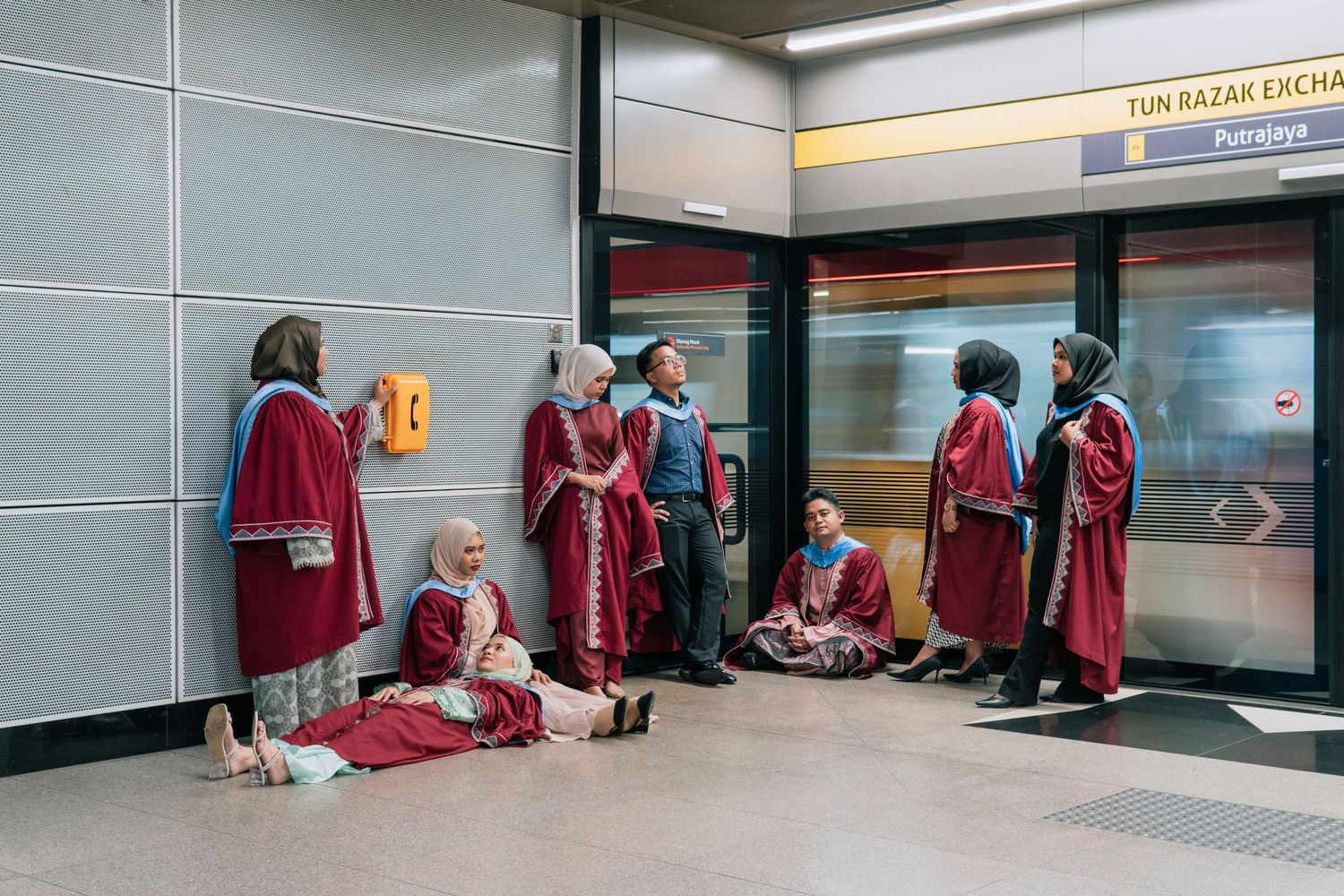 Students in red graduation gowns resting in a subway station corridor.