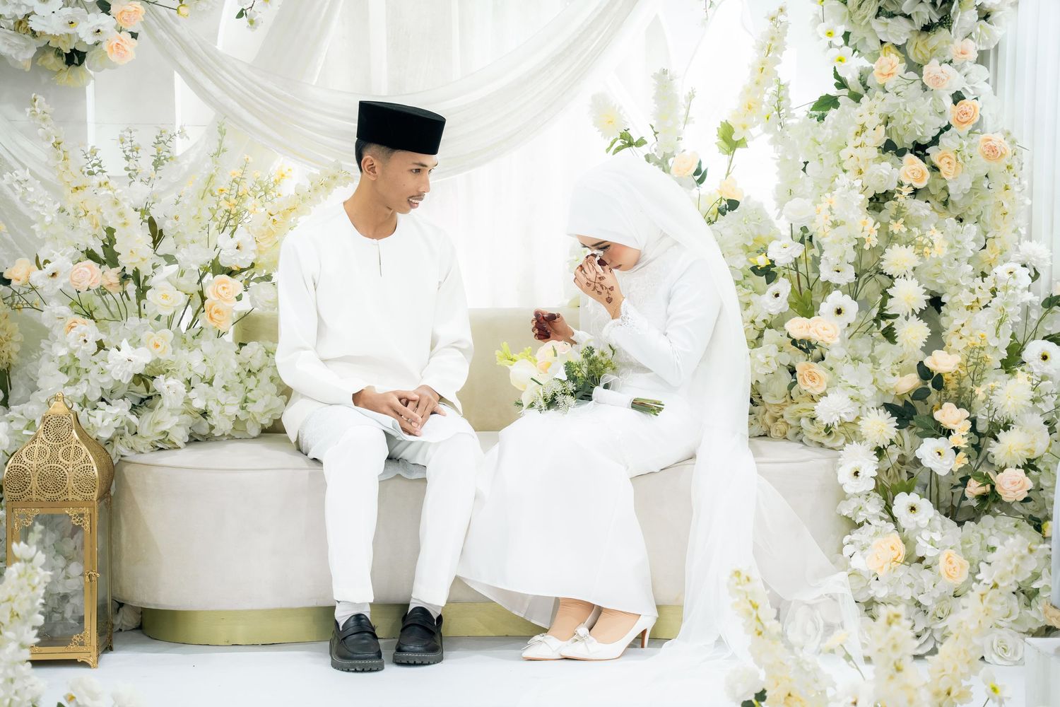 Traditional Malay wedding ceremony with couple in white attire surrounded by elegant white and blue floral decor.