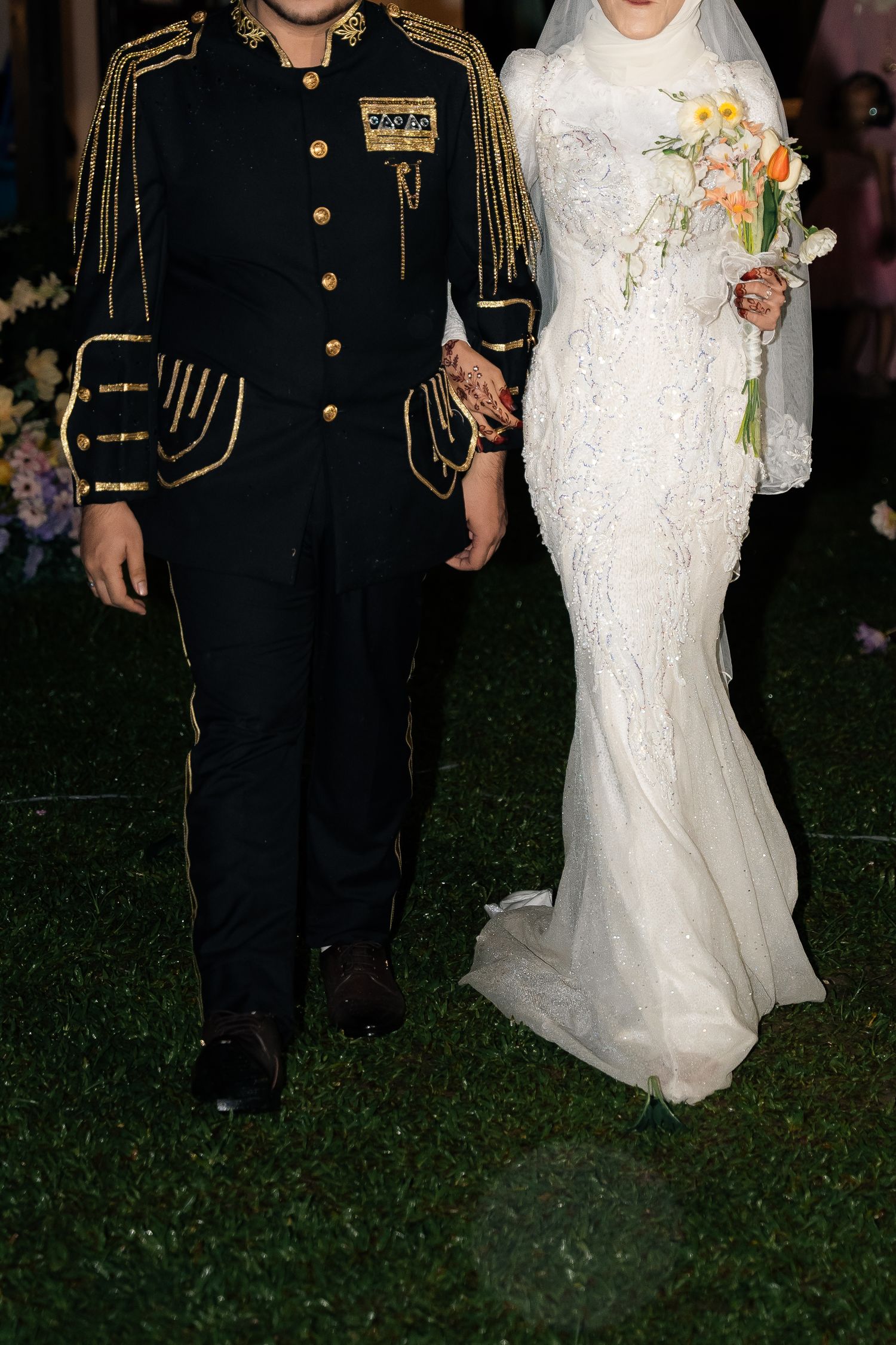 Military dress uniform and lace wedding gown couple walk together on grass during evening reception.