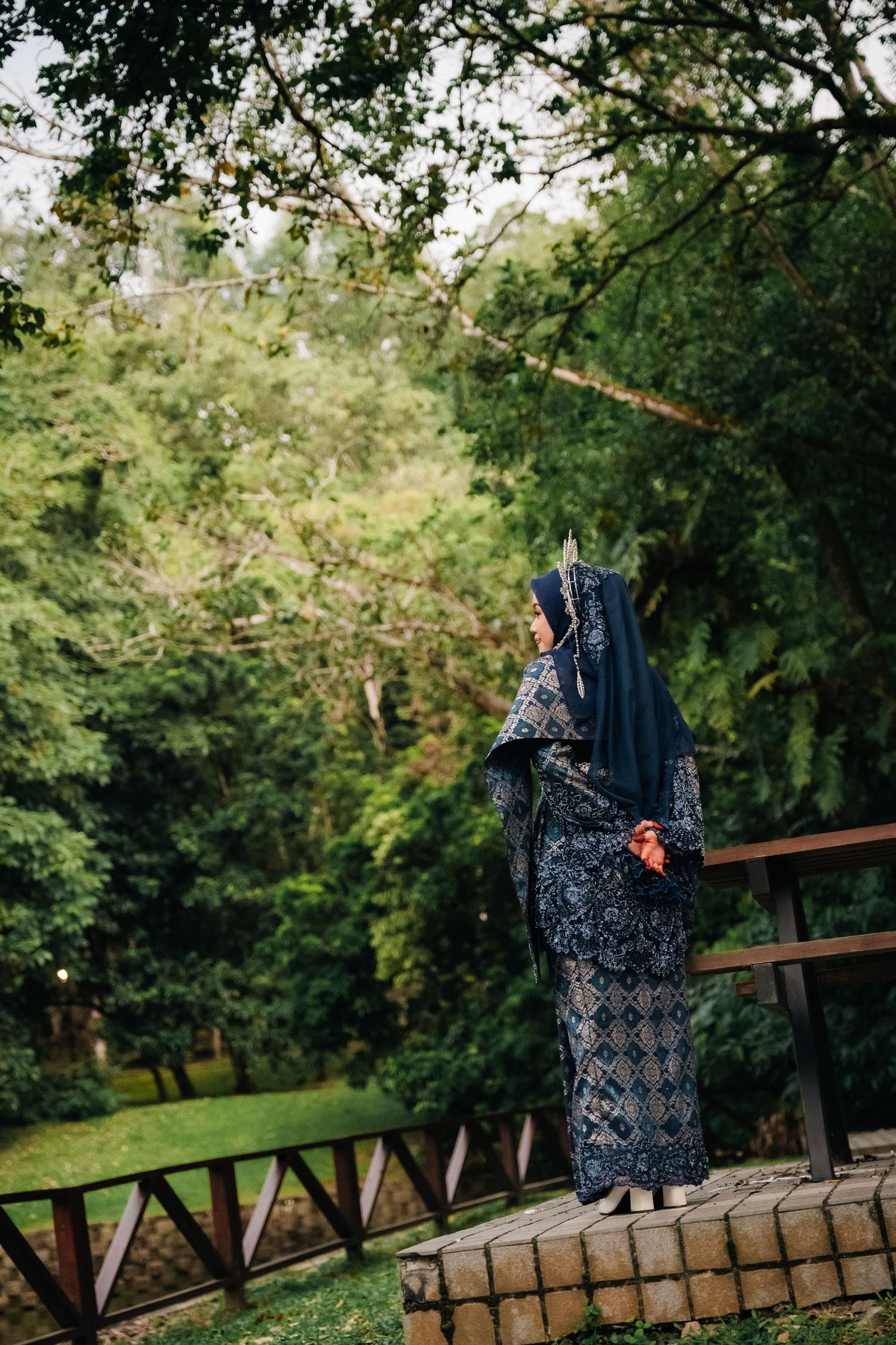 Person in a blue traditional dress and hijab standing on stone wall in lush green garden setting.