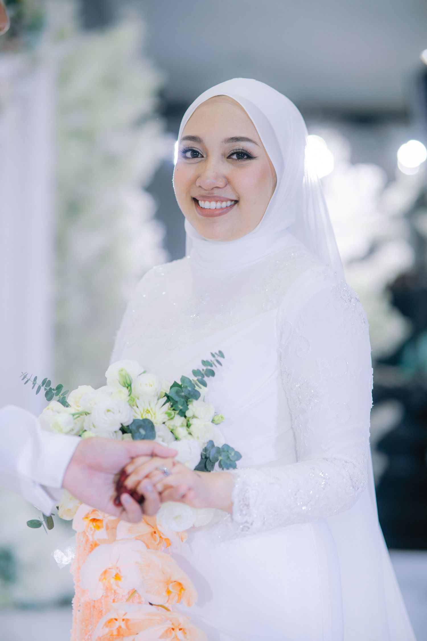 Beautiful bride in white hijab and modest dress holds delicate bouquet of white flowers and eucalyptus at wedding ceremony.