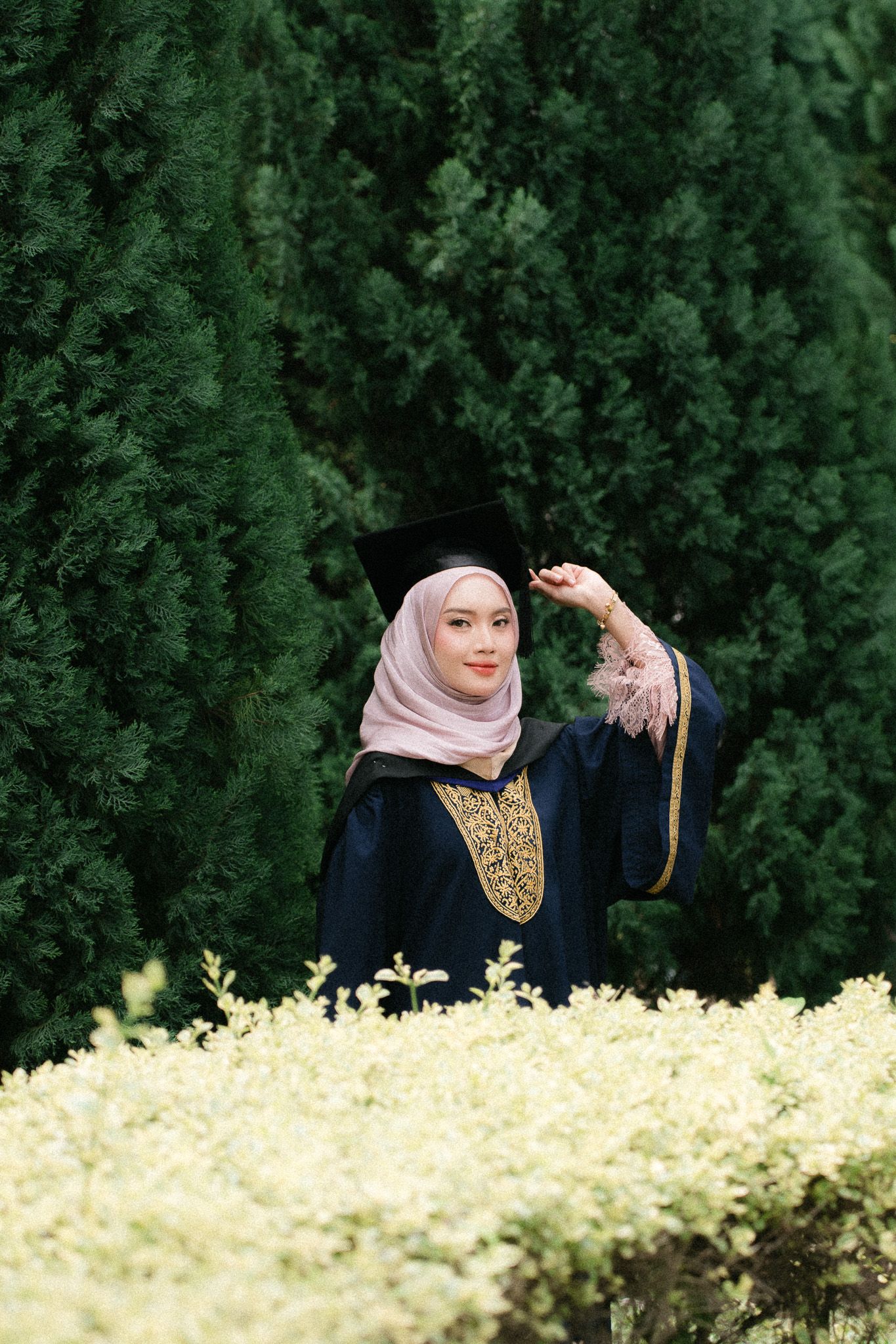 Muslim woman in hijab and graduation gown holding mortarboard cap outdoors among green trees.