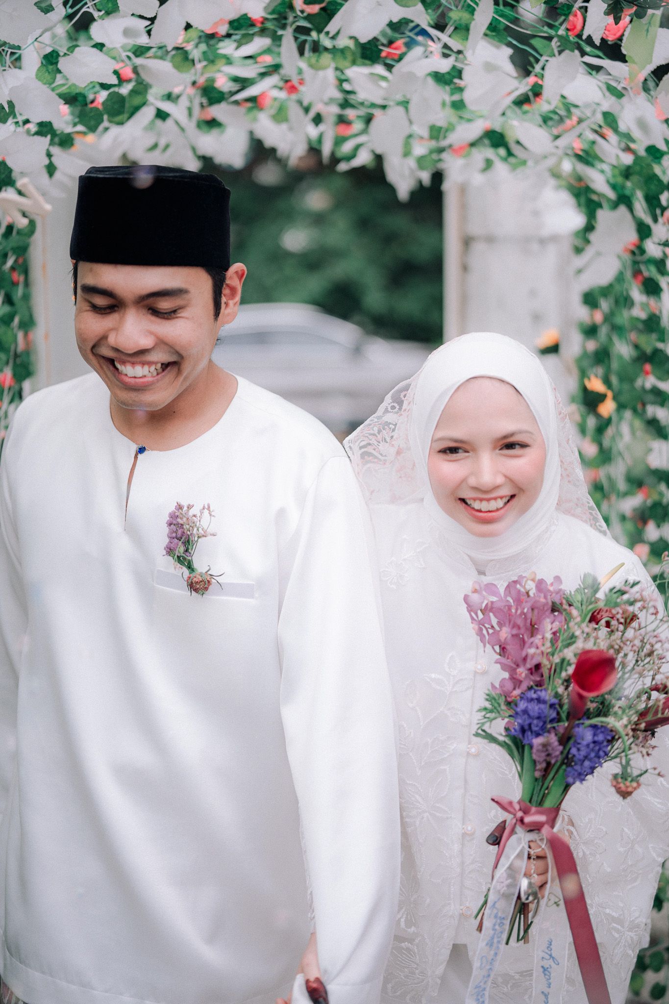 A smiling couple in traditional Malay wedding attire, groom in white baju melayu and songkok, bride in white hijab holding flowers.