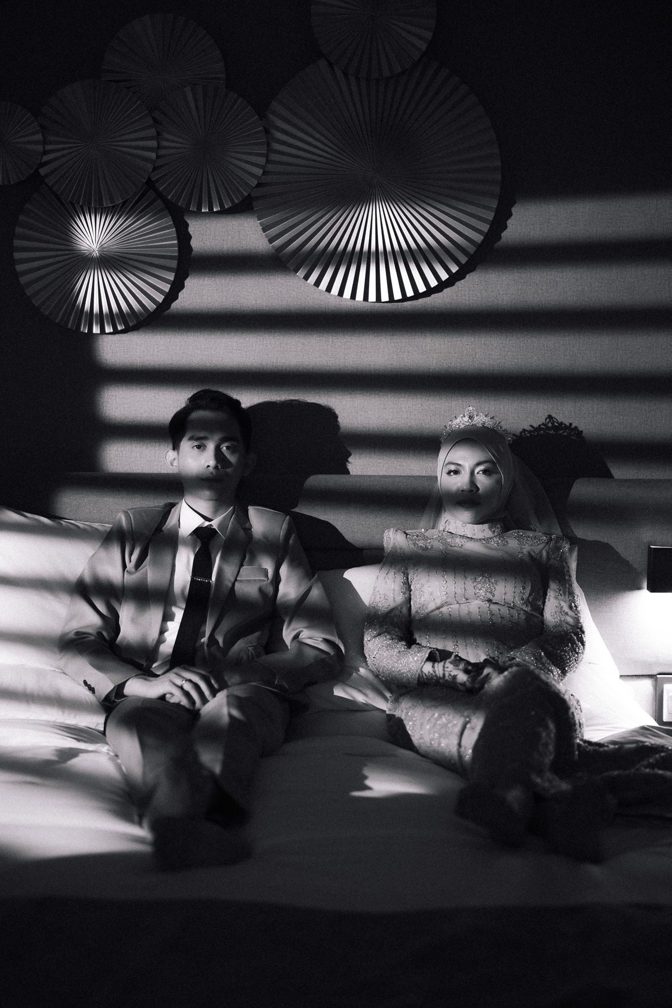 Black and white photo of a couple in wedding attire sitting on a bed under decorative paper fans in dramatic lighting.