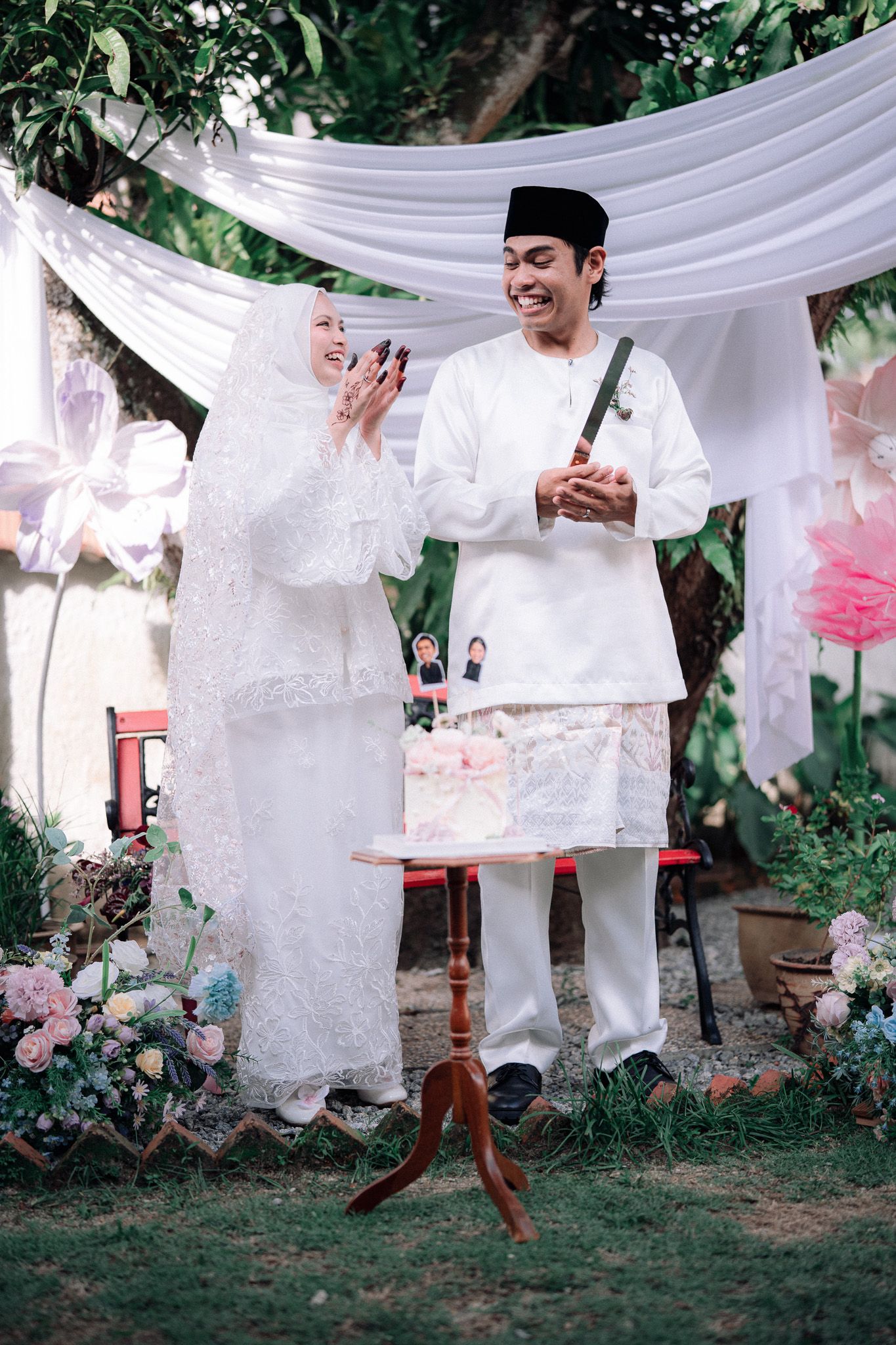 Muslim couple in white traditional attire cutting wedding cake outdoors, surrounded by floral decorations.