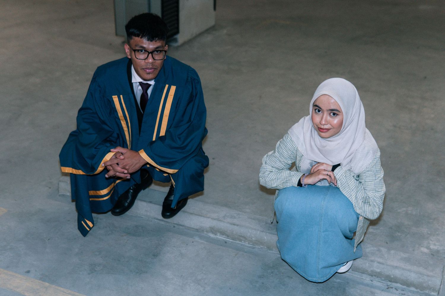 Two graduates in formal attire sit on the floor in a modern building hallway.