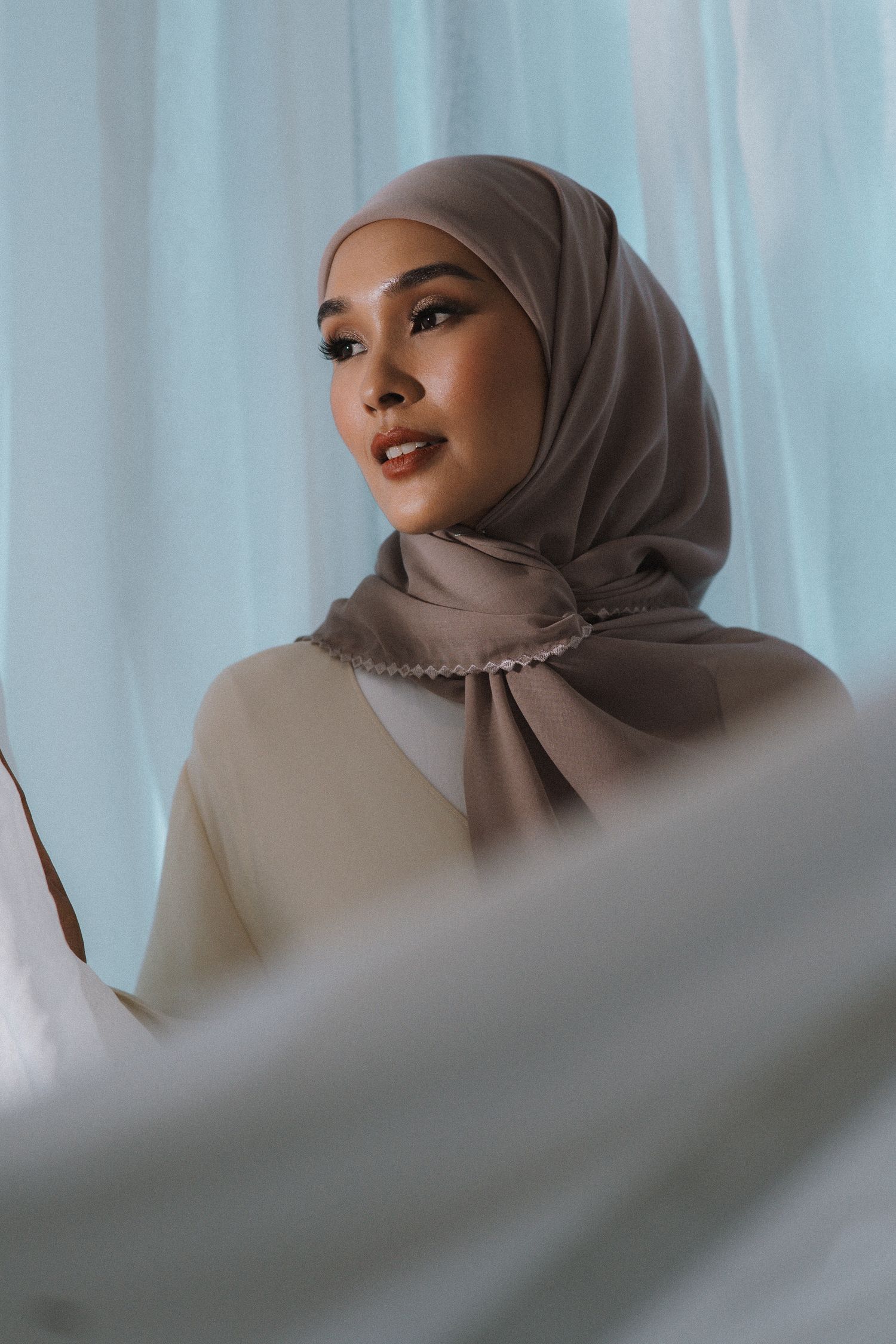 Portrait featuring taupe hijab and neutral-toned outfit against soft blue backdrop.