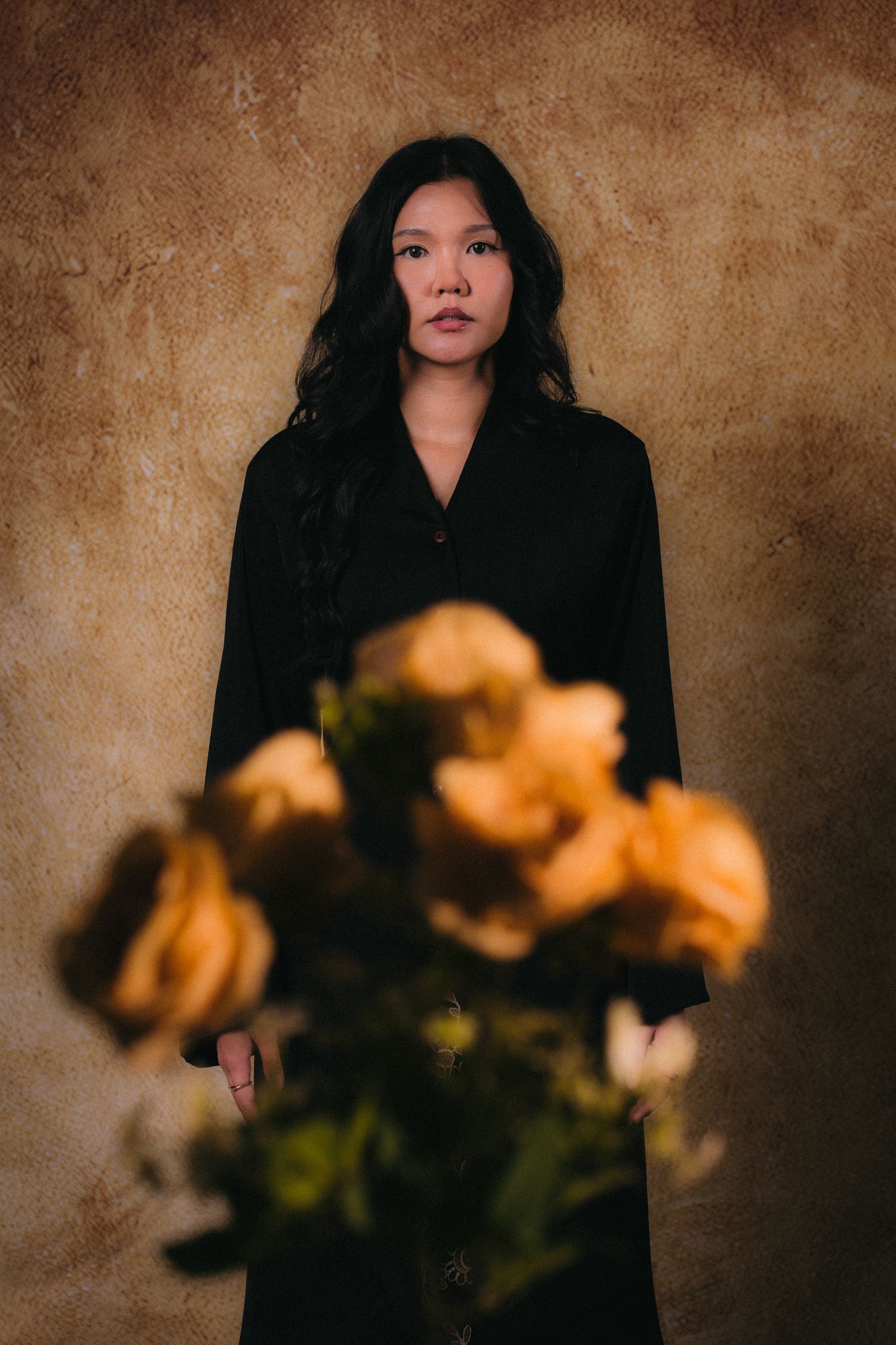 A moody portrait with orange roses in foreground and figure in black against textured brown wall.