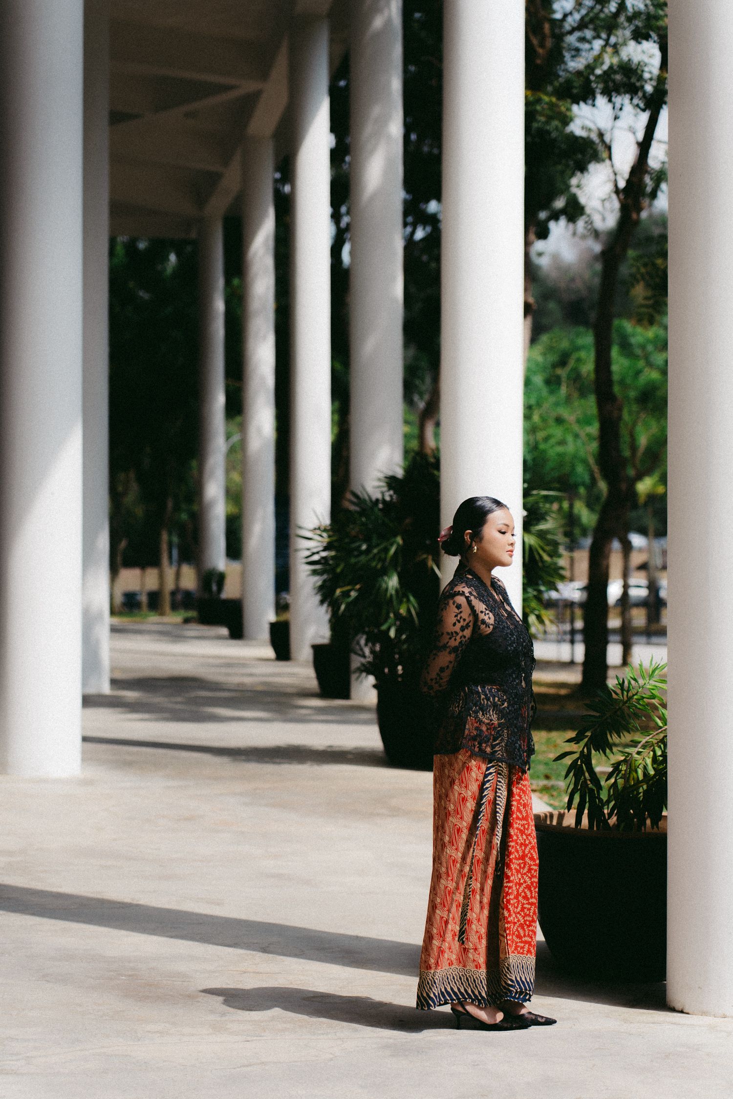 Figure photographed in elegant lace and floral dress stands between white columns in an outdoor architectural setting.