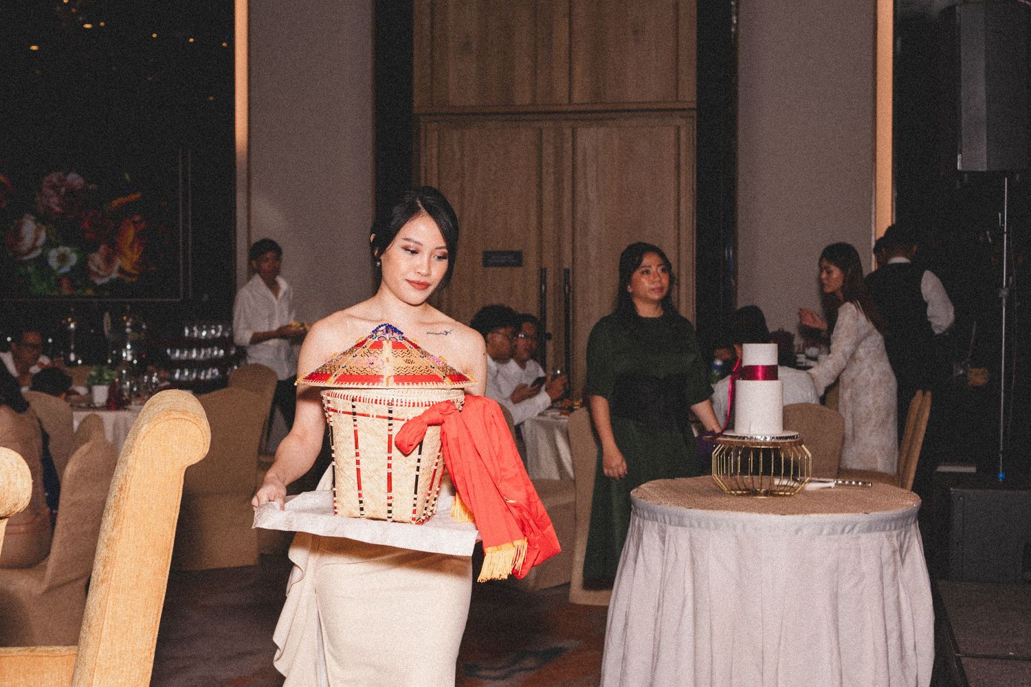 A waitress in a white uniform carries plates at an indoor event with round tables and guests in the background.