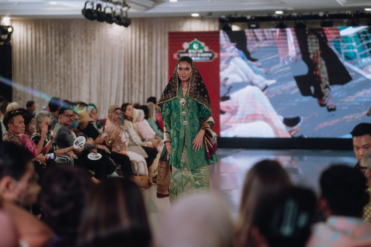 Fashion show audience watches model in emerald green traditional attire on catwalk.