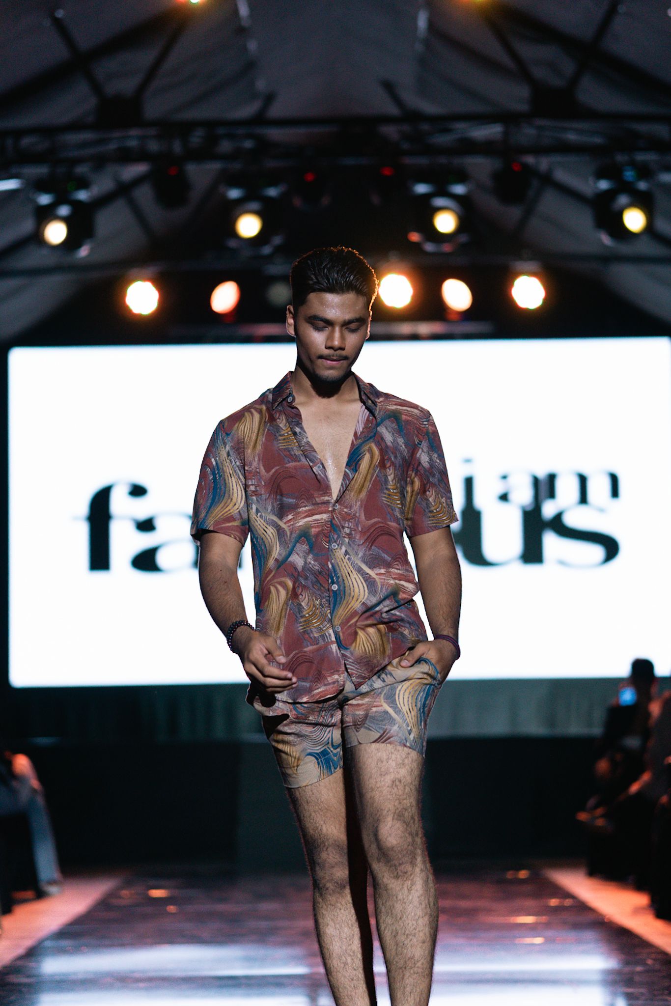Model walks runway in colorful printed shorts and matching shirt under dramatic lighting.
