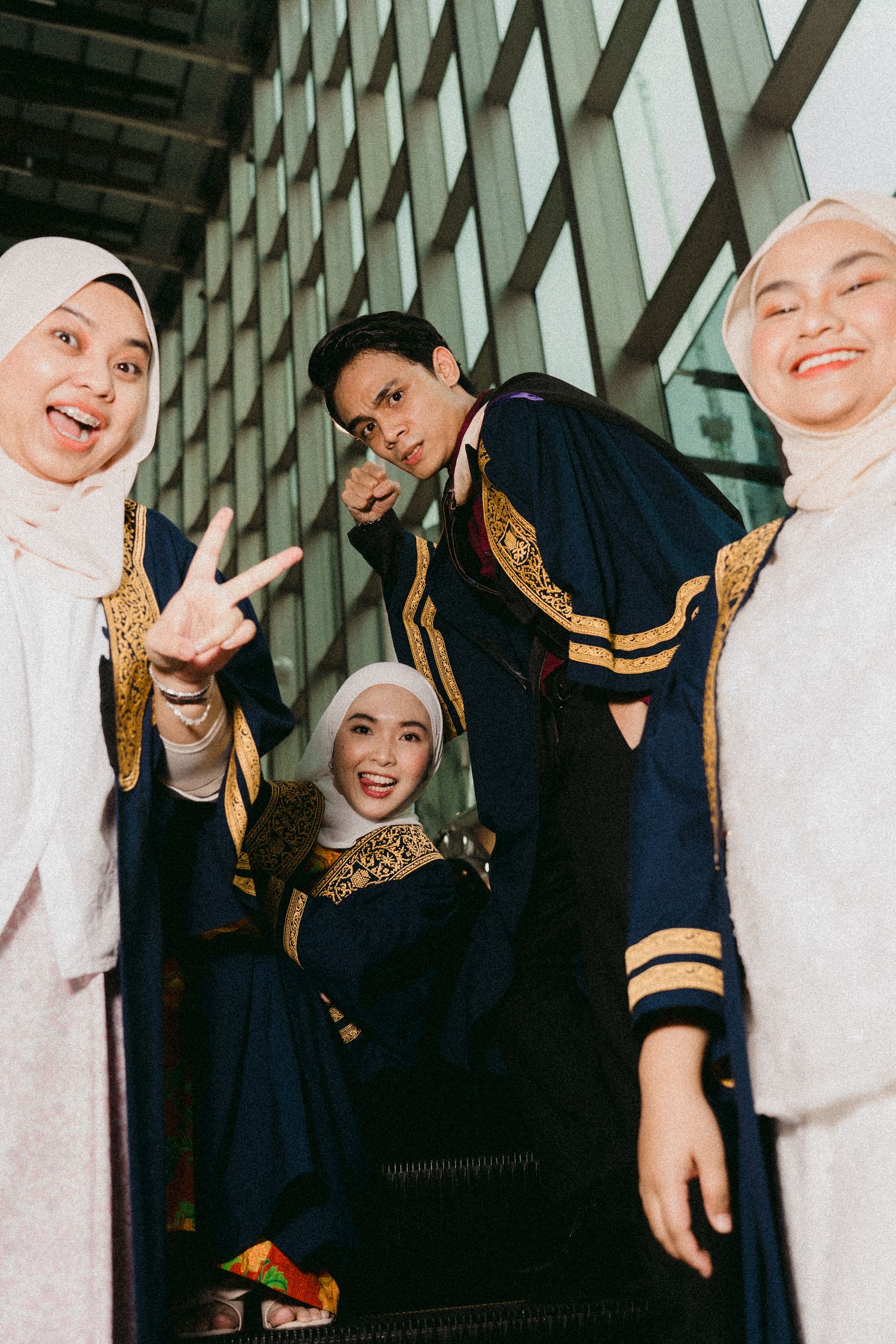 Graduates in ceremonial dress pose playfully near a modern glass building facade.