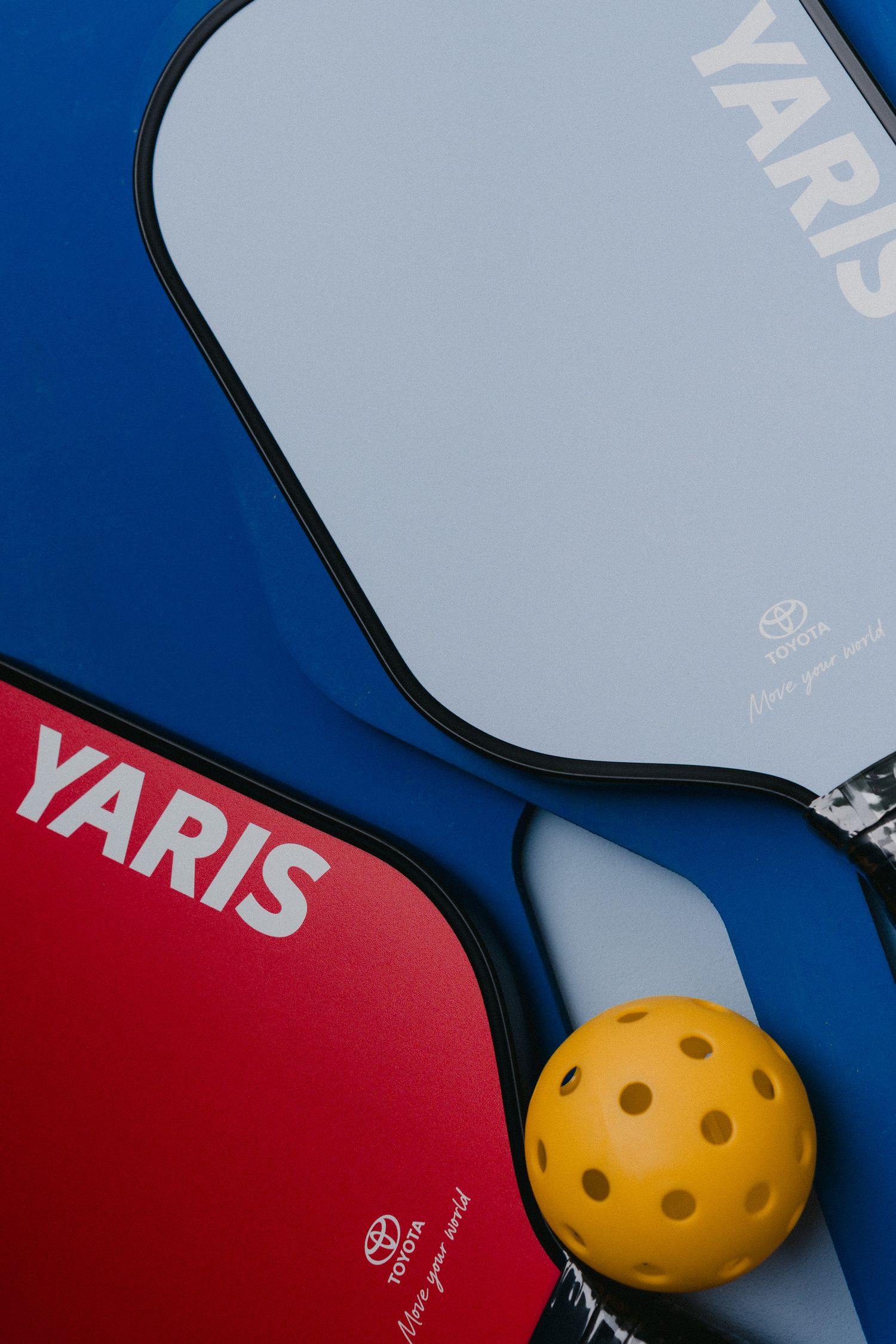 Close-up view of pickleball paddles and yellow ball against blue and red background showing equipment details.