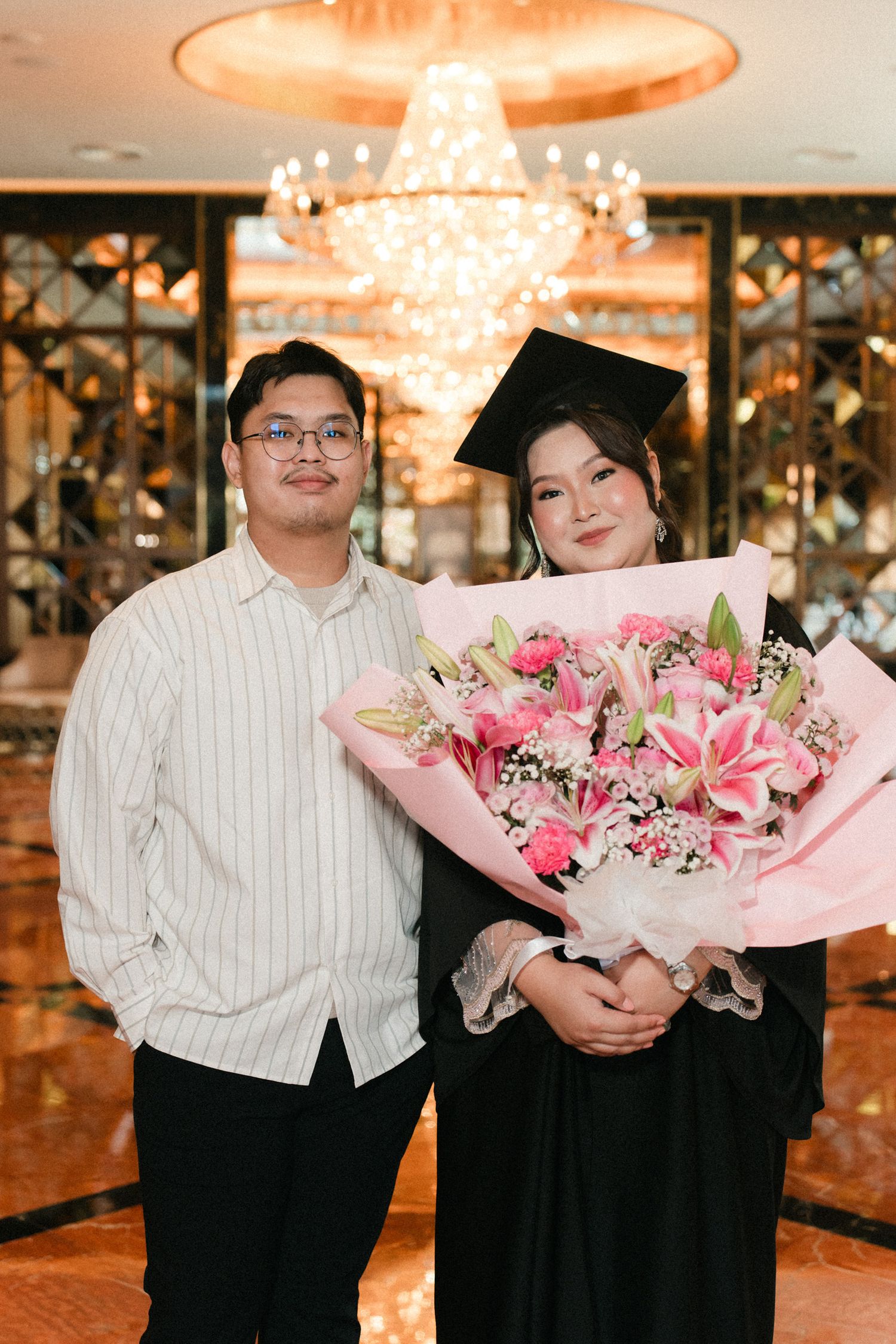 A couple stands together in a formal setting with pink floral bouquets under warm chandelier lighting.