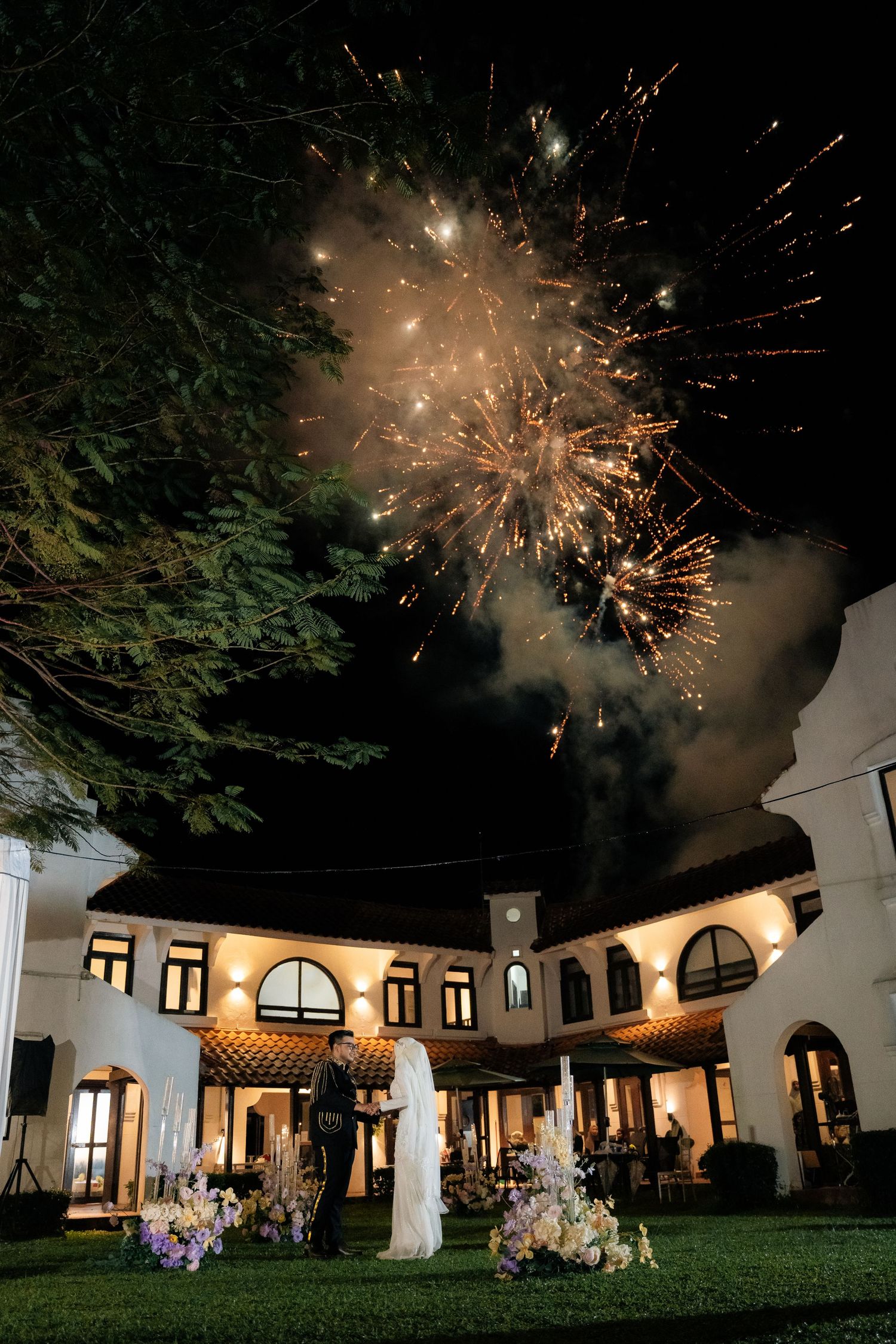 Fireworks explode over an elegant white villa with arched windows and columns during a nighttime celebration on a manicured lawn.