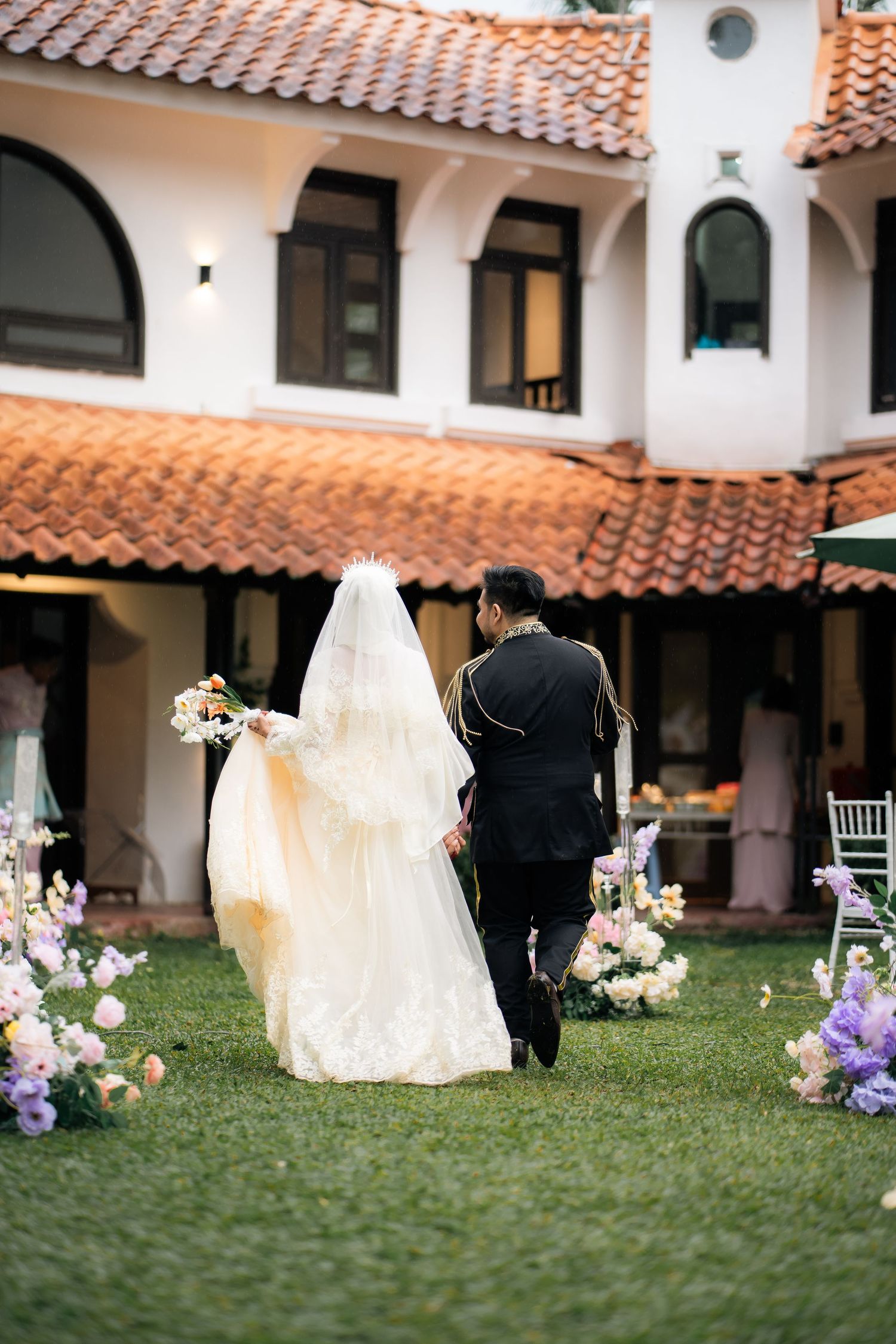 A bride and groom walk together on a grassy lawn with Spanish-style architecture and floral decorations in the background.