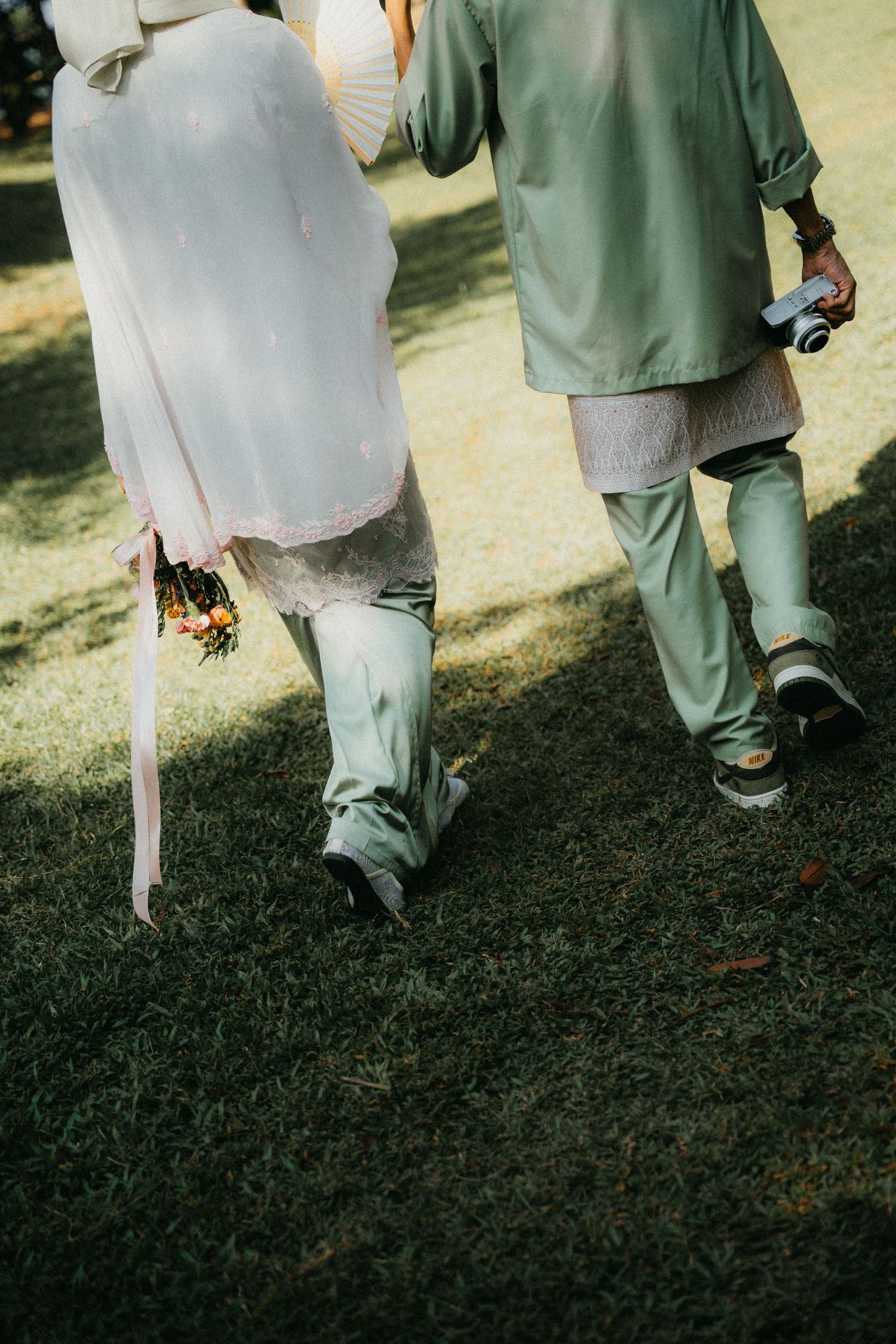 Two people walk on grass outdoors, one wearing green and one wearing light colored formal attire.