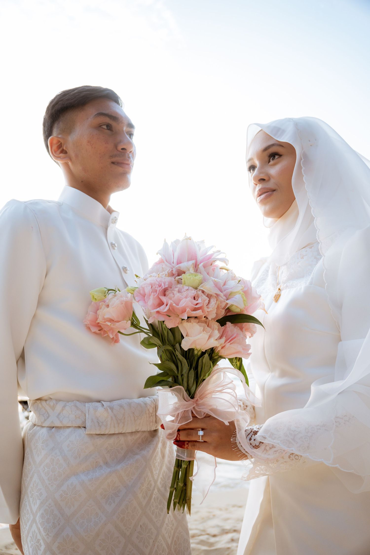 Bride and groom in traditional white Muslim wedding attire pose together with pink floral bouquet against bright sky.
