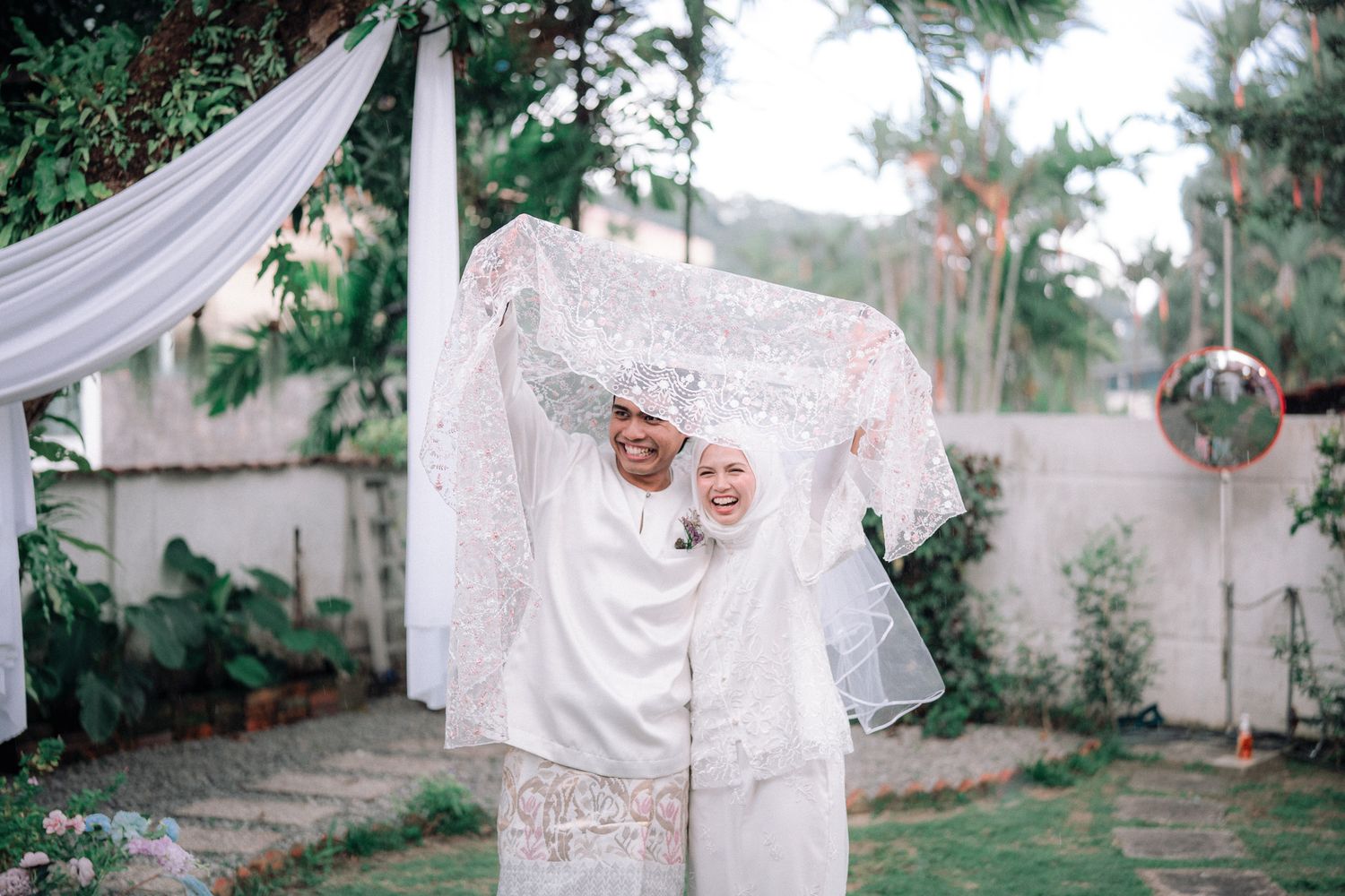 Muslim couple in white wedding attire sharing a romantic moment under lace veil in a garden setting.