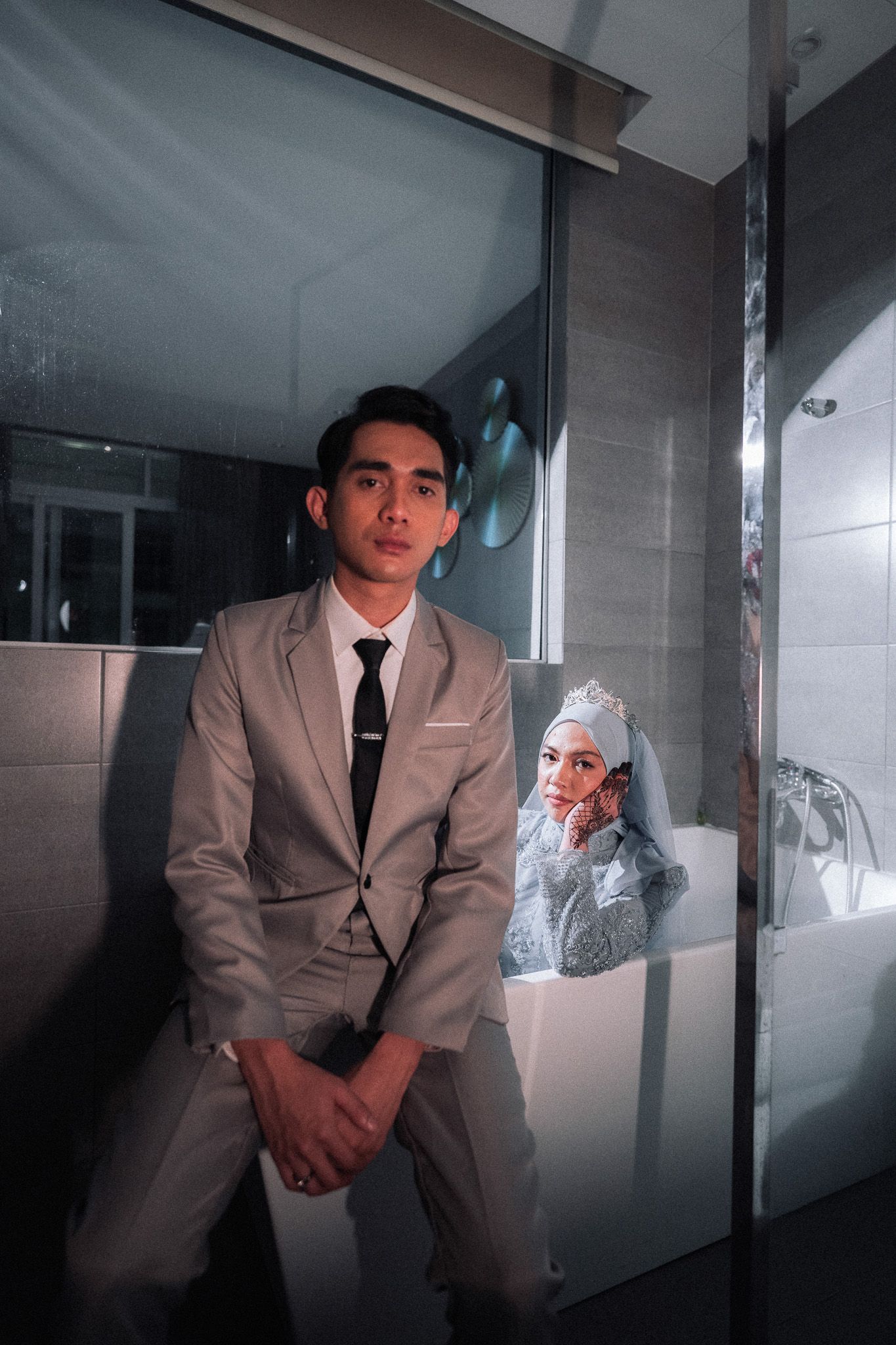 Groom in gray suit sits by glass door while bride in hijab poses in bathtub during wedding photoshoot.