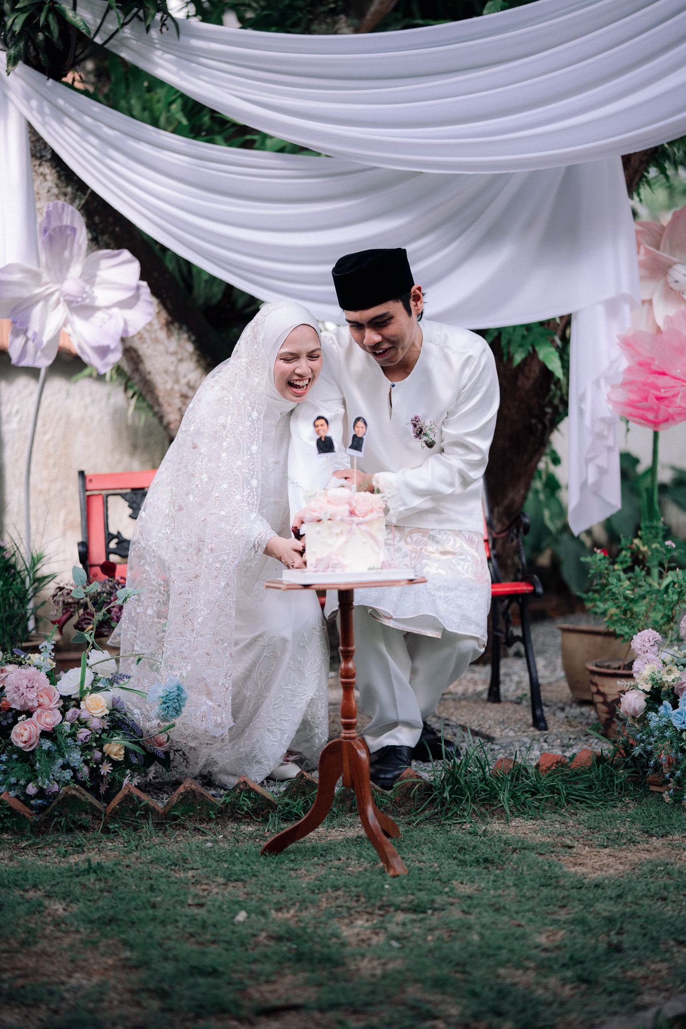 Muslim bride and groom cutting wedding cake outdoors in traditional white attire under draped fabric canopy.