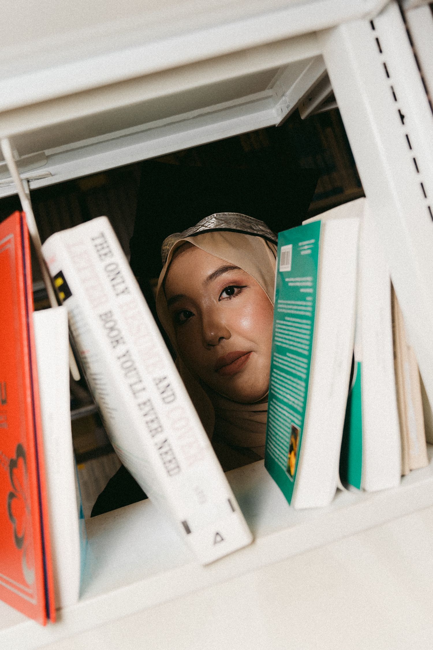 A creative portrait through a bookshelf shows someone peeking between colorful book spines on white shelving.