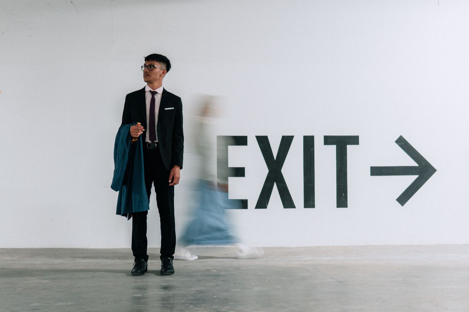 A figure in a business suit stands near an exit sign on a minimalist white wall.