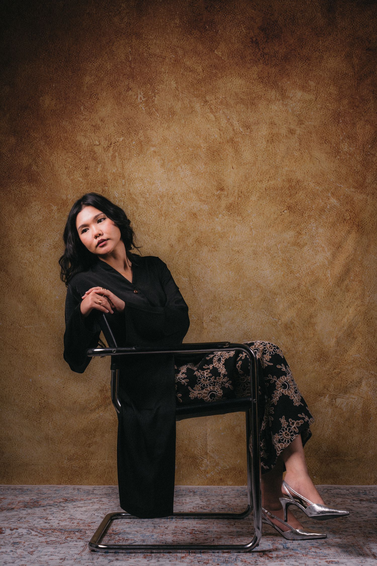 A dramatic portrait of a person in black clothing seated on a metal chair against a brown backdrop.