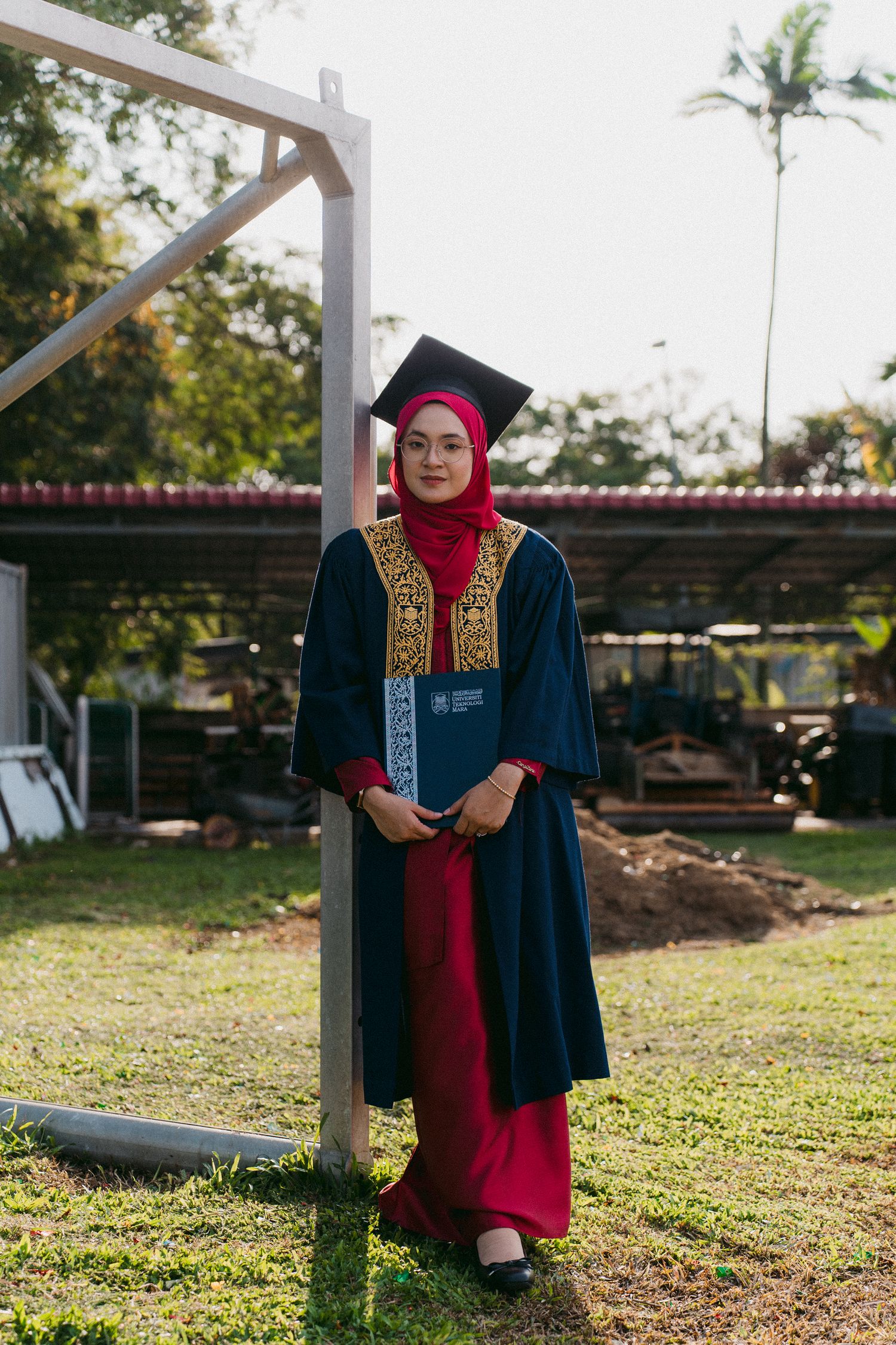 Graduate in navy and red academic regalia stands beside outdoor pole in garden setting.