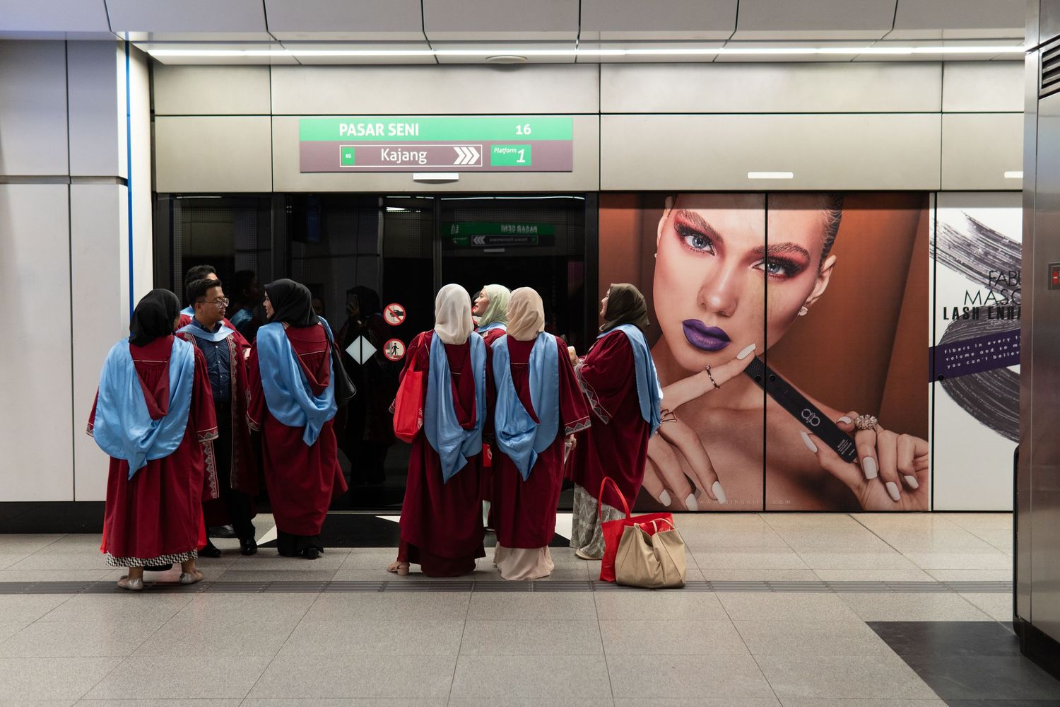 Students in red graduation robes walking past a fashion advertisement in a subway station.