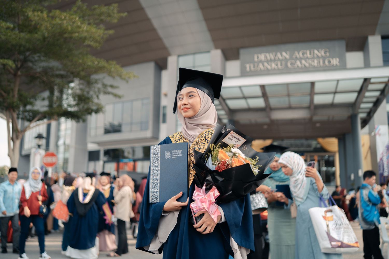 Graduate celebrating outside campus building holding colorful bouquet.