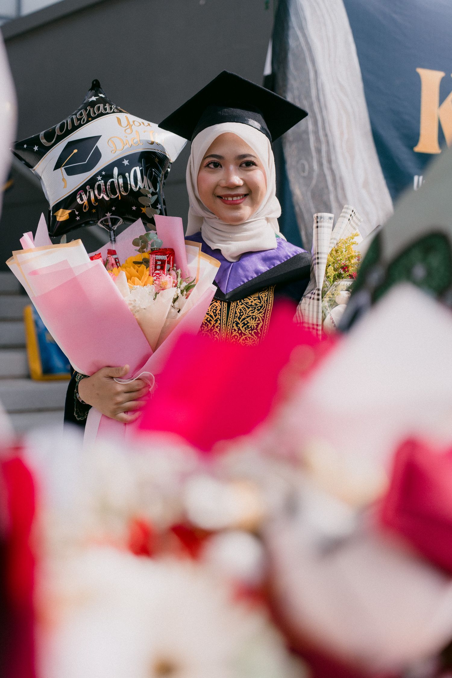 Graduates in traditional academic regalia and hijabs hold colorful bouquets during a graduation ceremony celebration.