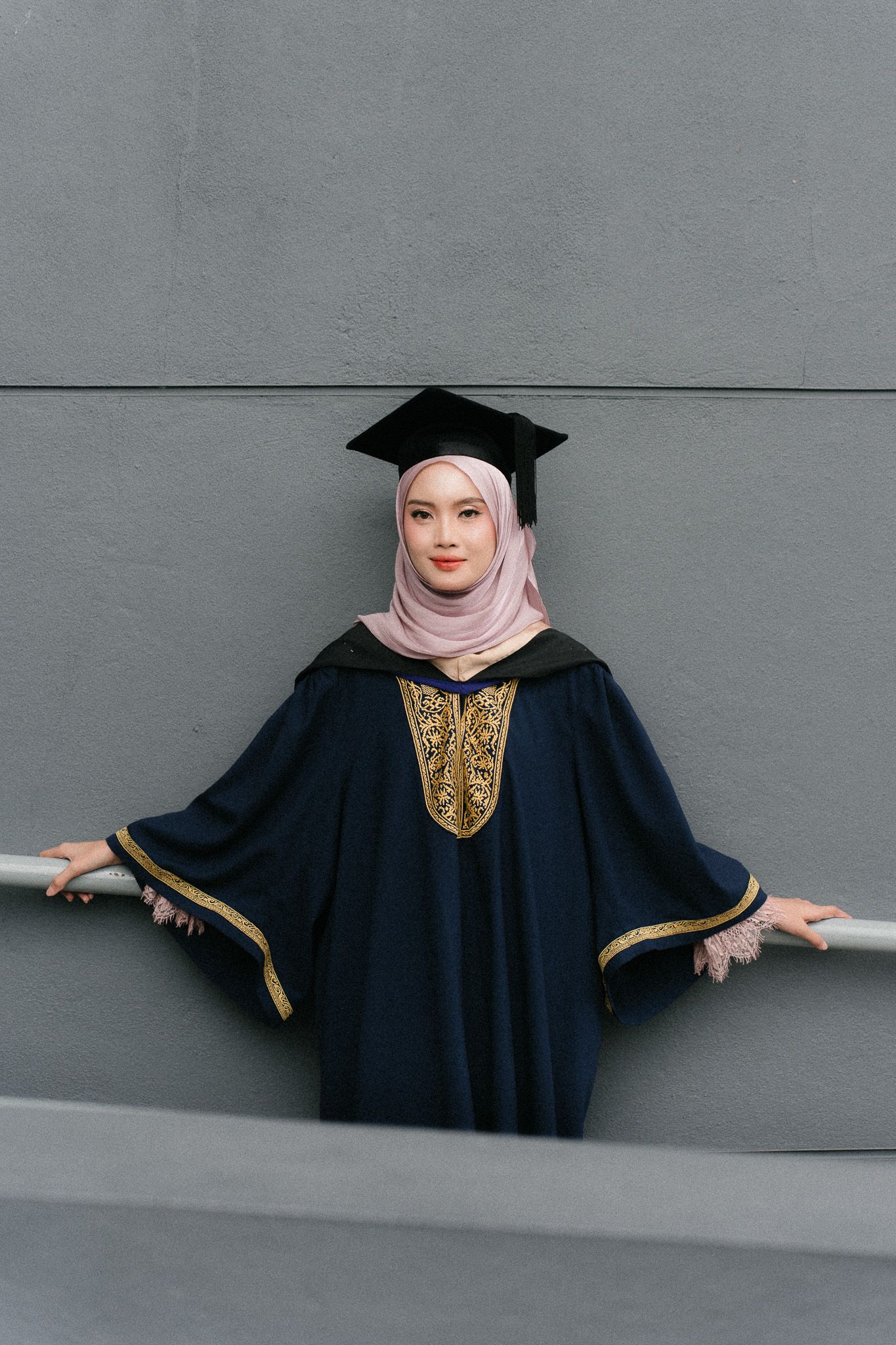 Young Muslim woman in navy graduation gown and mortarboard cap with pink hijab posing against gray wall.