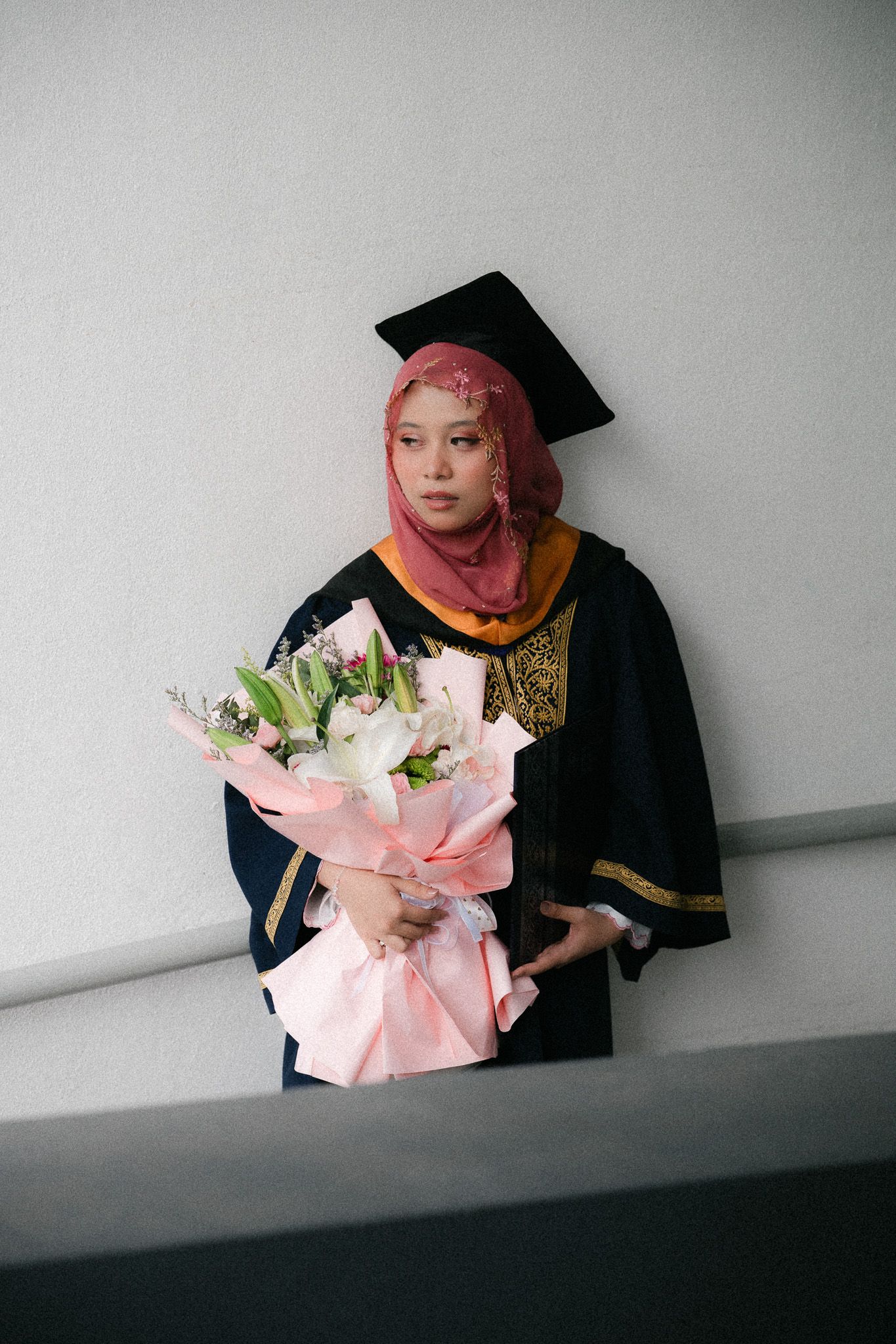 Graduate in black gown and pink hijab holding a bouquet of white and pink flowers at graduation ceremony.