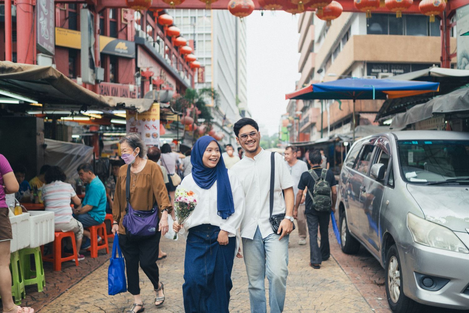 Two people walking through a busy street market with vendors and parked cars.