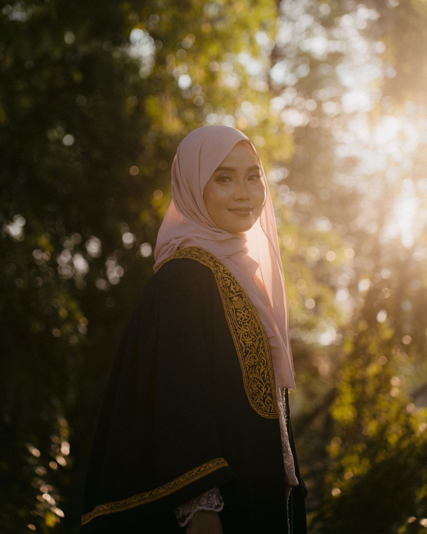 A person in a pink hijab stands in golden sunlight filtering through trees in a backlit outdoor portrait.