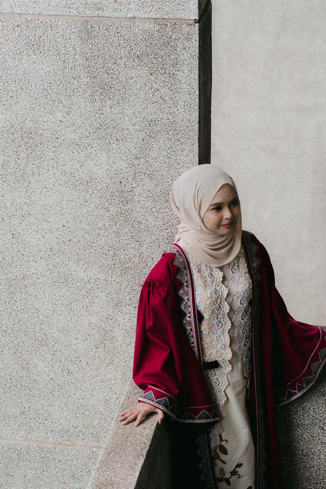 A young woman wearing a cream hijab and burgundy embroidered abaya poses gracefully against a textured stone wall.