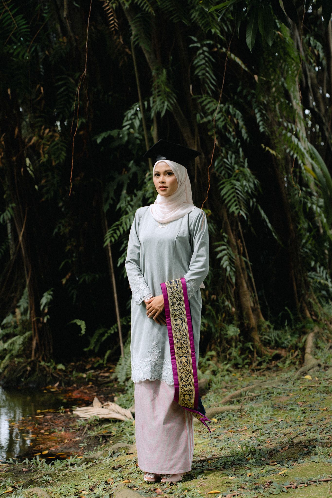 Muslim woman in hijab and traditional baju kurung standing in lush tropical forest setting.
