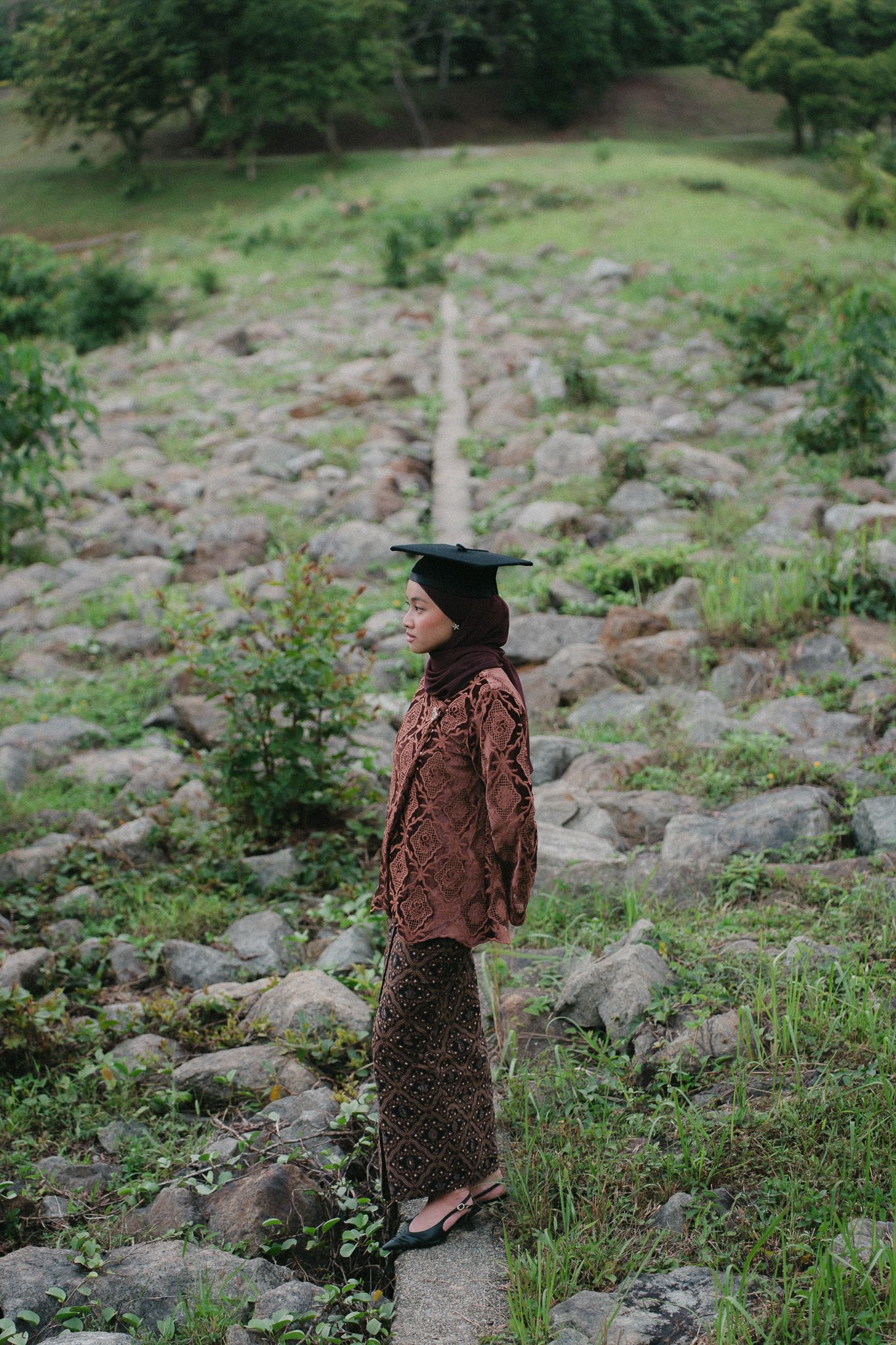 A woman in traditional batik attire and graduation cap stands on rocky terrain surrounded by lush greenery.
