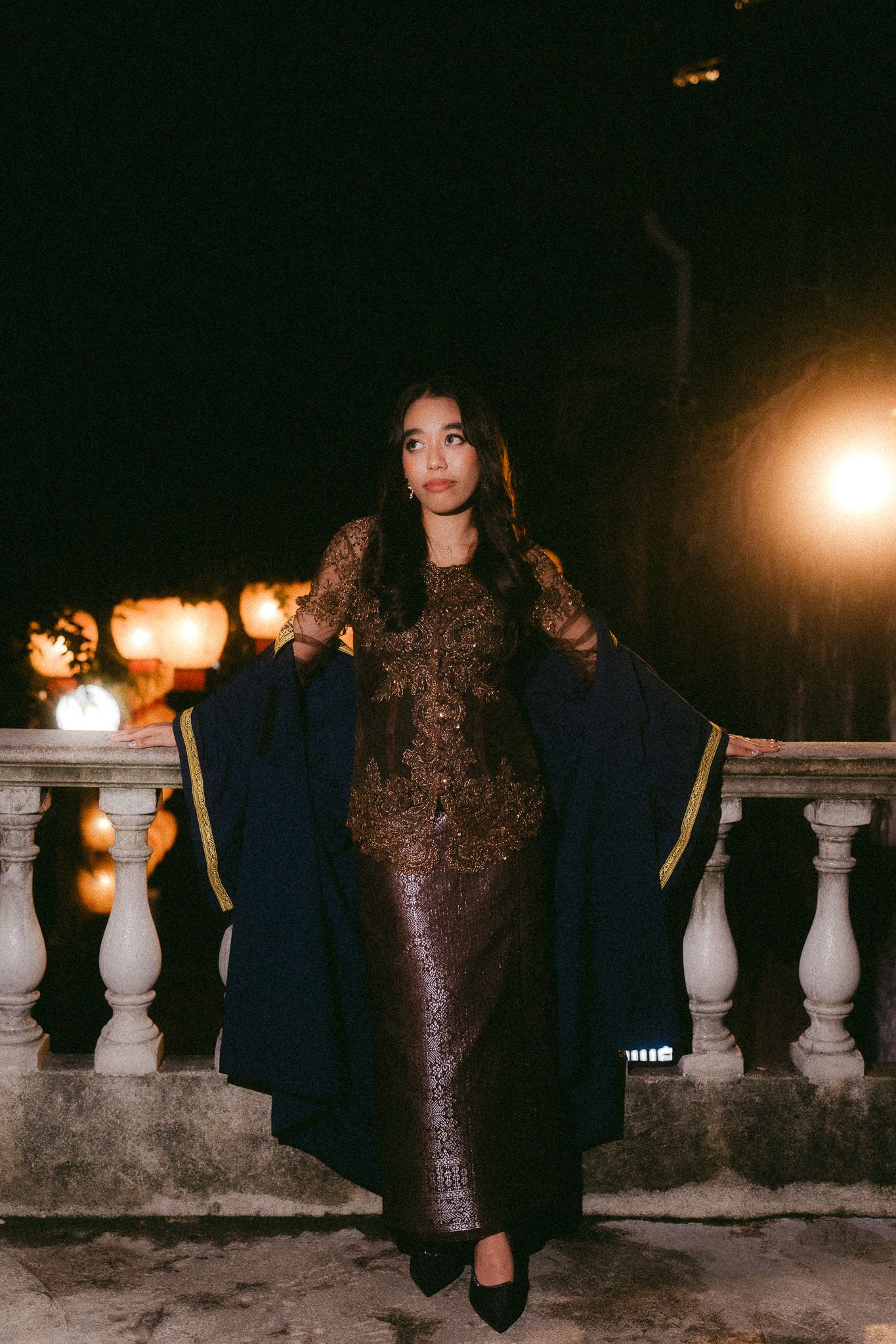 A woman in a gold embroidered gown holds a navy coat on a stone balcony at night.